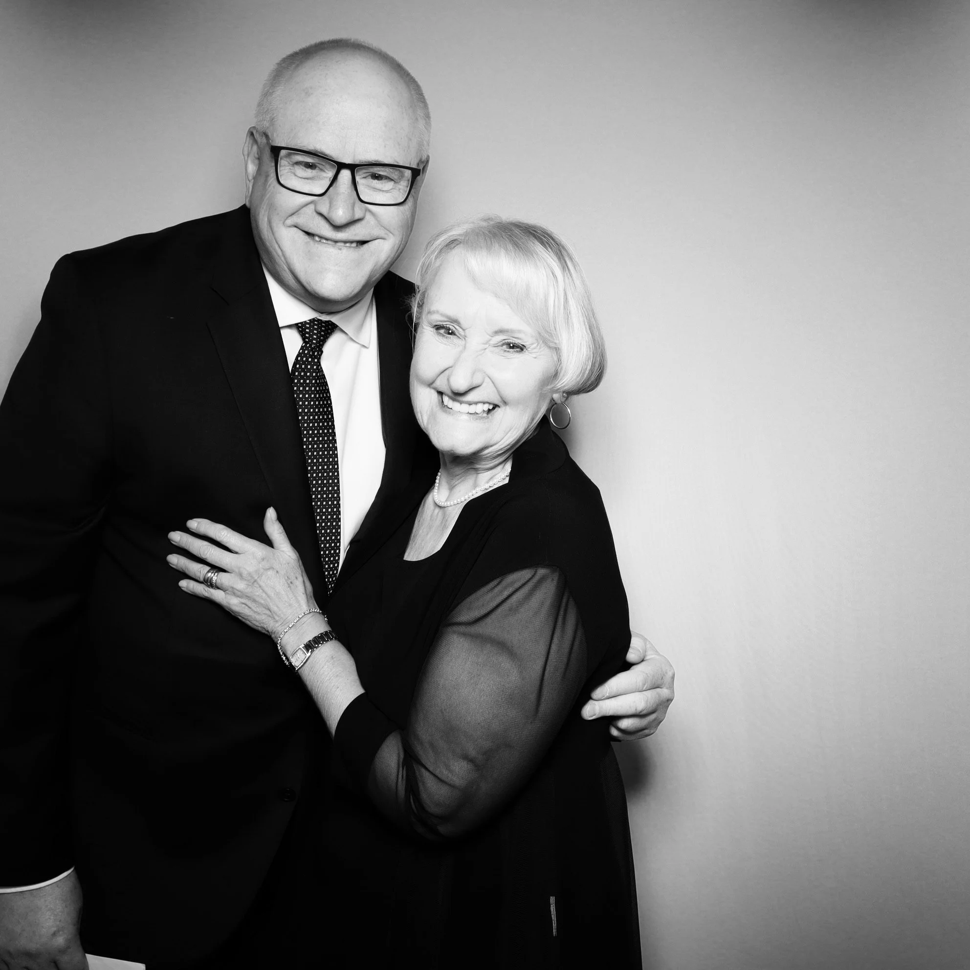 Black and white photo of a smiling elderly woman and a middle-aged man hugging each other, both wearing dark clothing, in front of a plain wall.