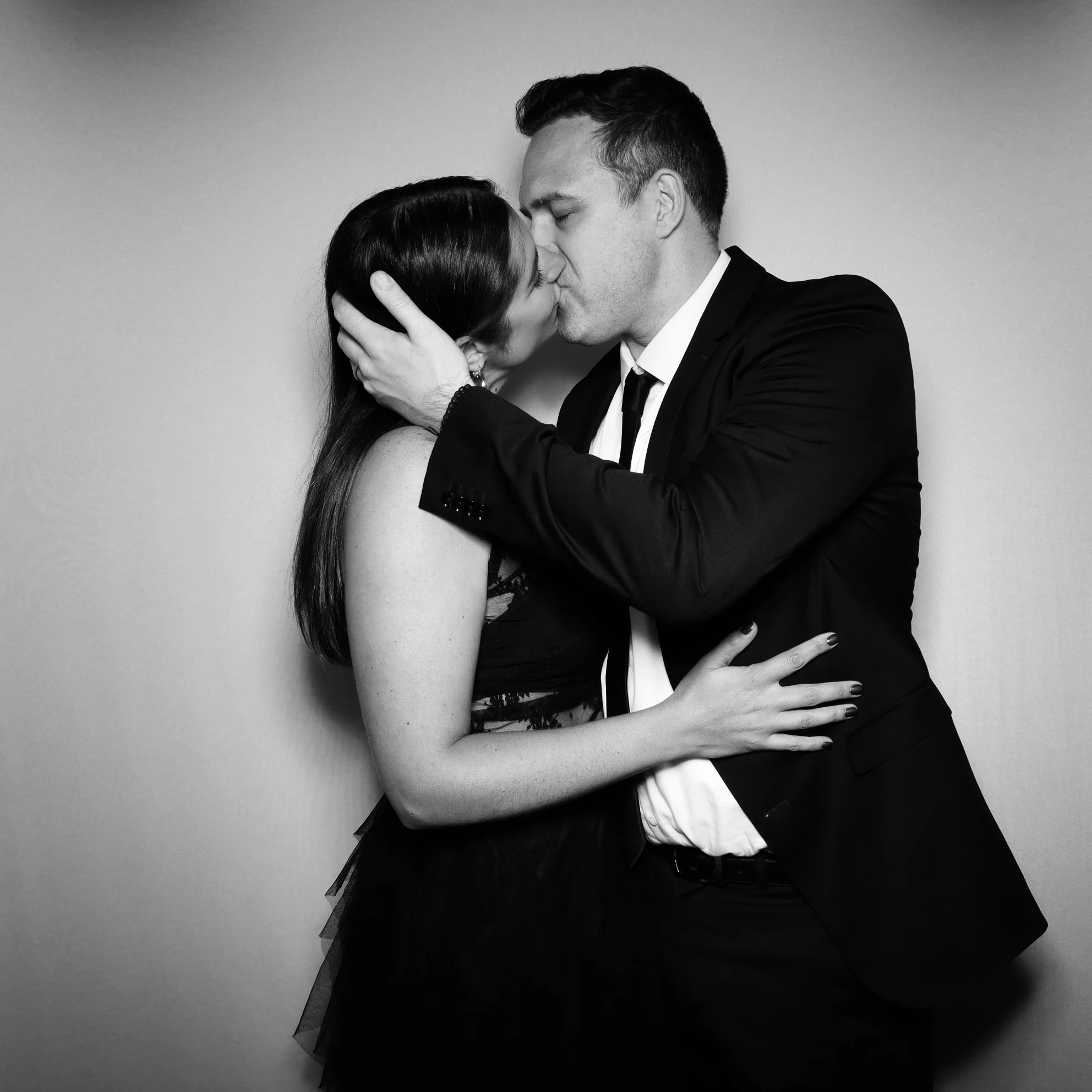 A black and white photo of a couple kissing. The woman has long dark hair and is wearing a dress, while the man is in a suit. They are close together, embracing and sharing an intimate moment.