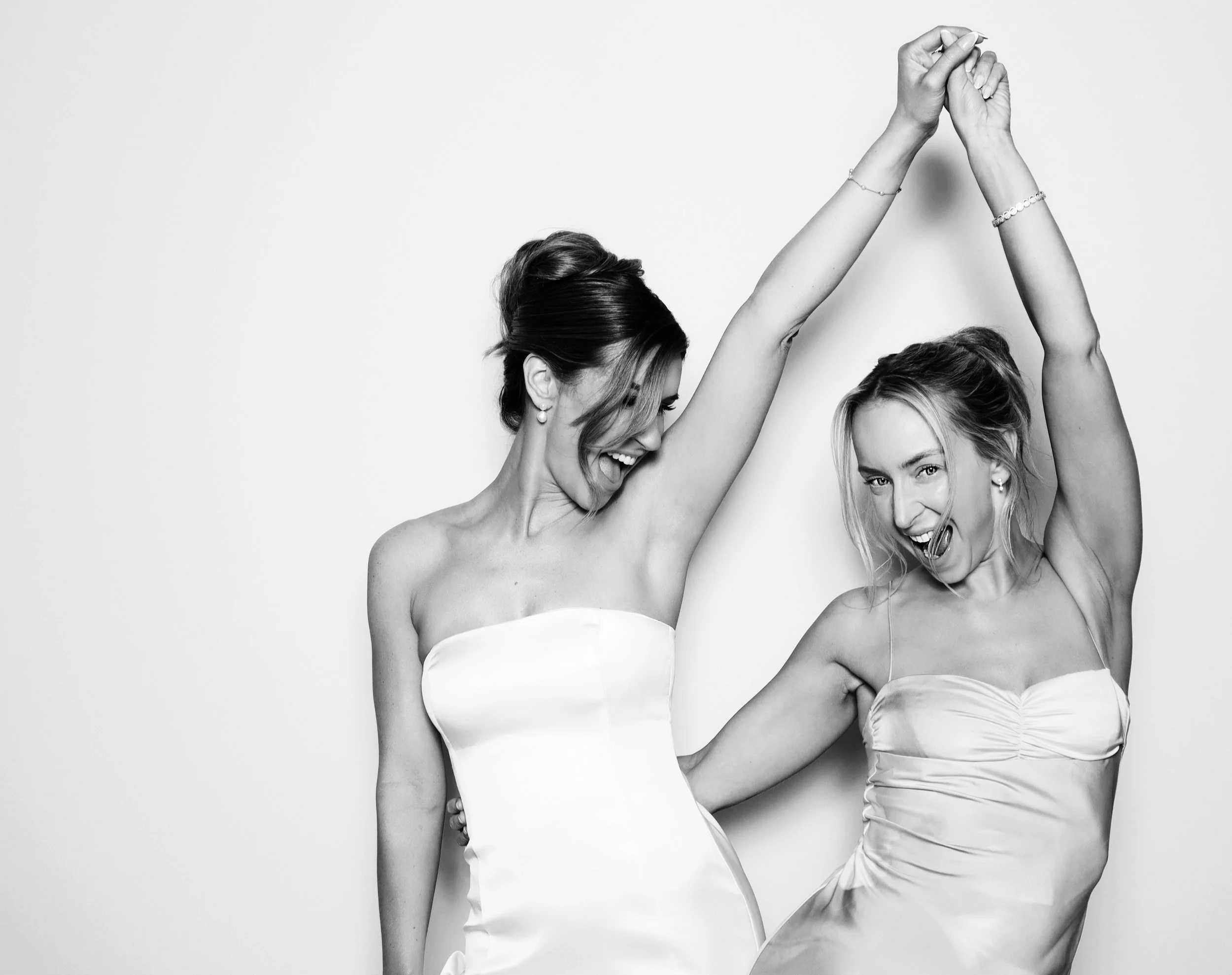 Two women in strapless dresses are joyfully dancing together, smiling and raising their hands in a celebratory manner.