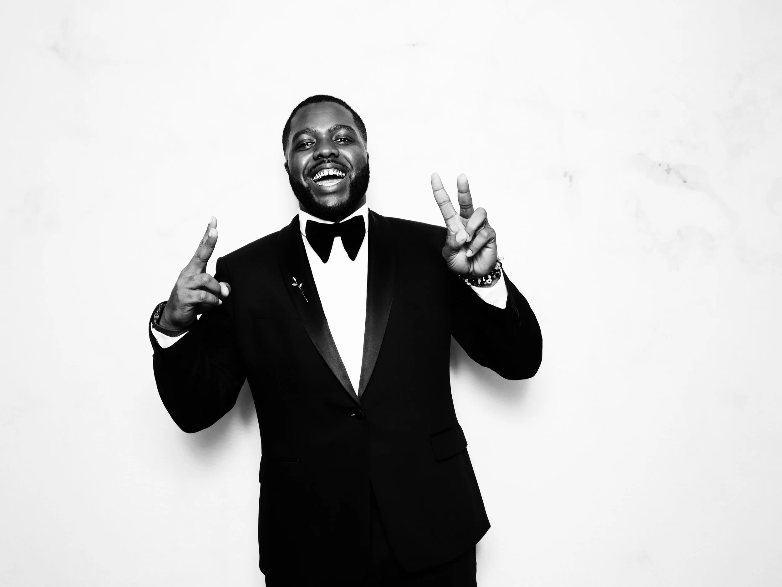 A man in a tuxedo smiling and making peace signs with both hands against a plain white background.