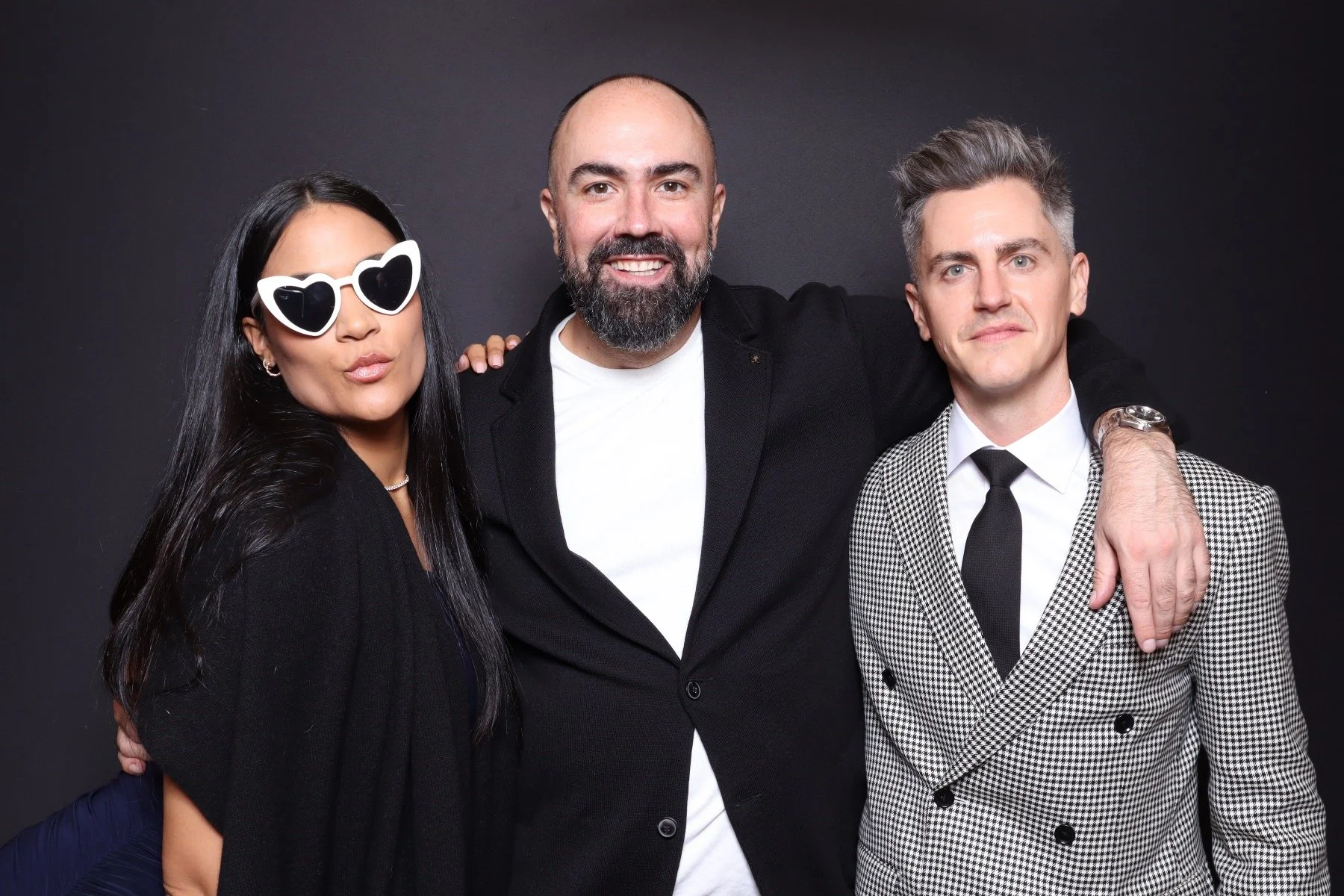 Three people posing together against a dark background. The woman on the left wears heart-shaped sunglasses and a black outfit. The man in the middle has a beard, a bald head, and wears a black blazer. The man on the right has gray hair, wears a chec