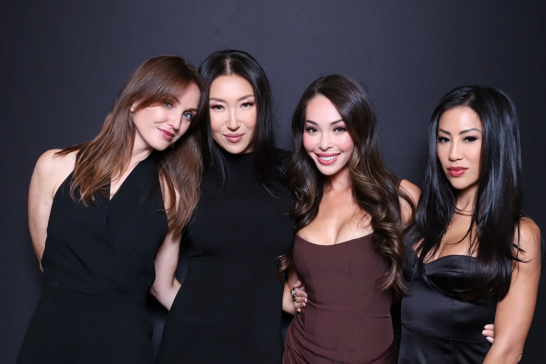 Four women posing together against a black background, smiling and dressed in stylish black and dark brown dresses.