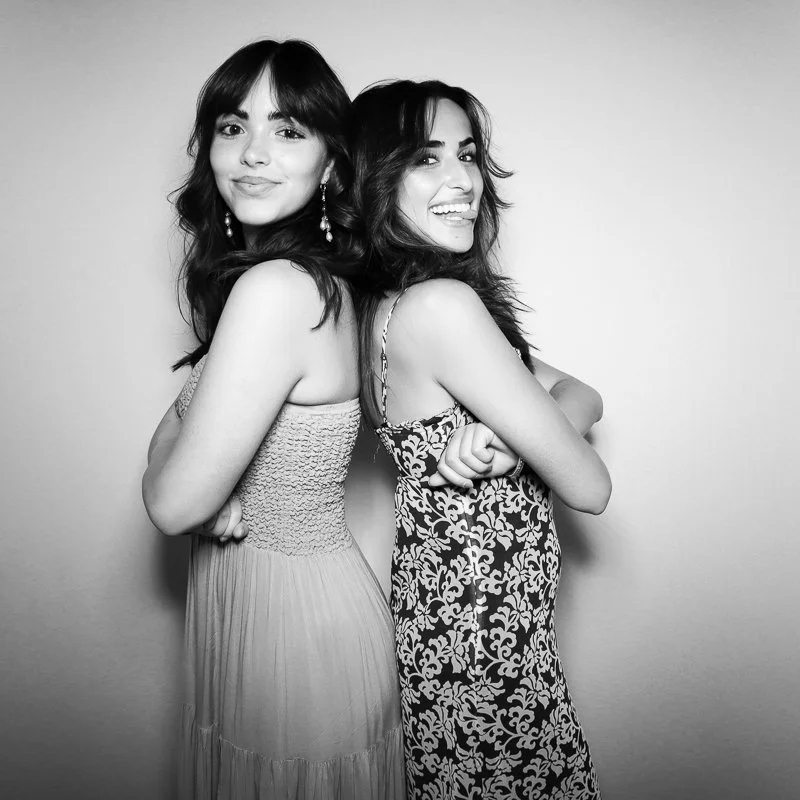 Two women standing back-to-back smiling at the camera, wearing fashionable dresses, in black and white