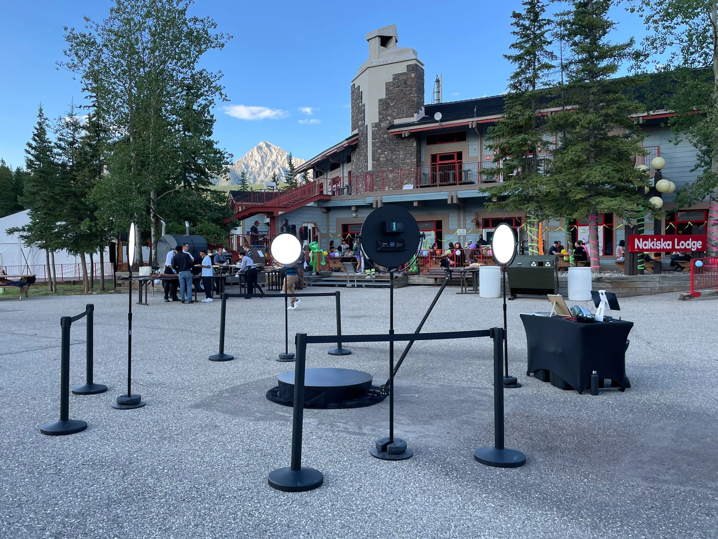 Outdoor event setup in front of Nakiska Lodge, with tables, chairs, and lighting equipment, surrounded by trees and mountains in the background.