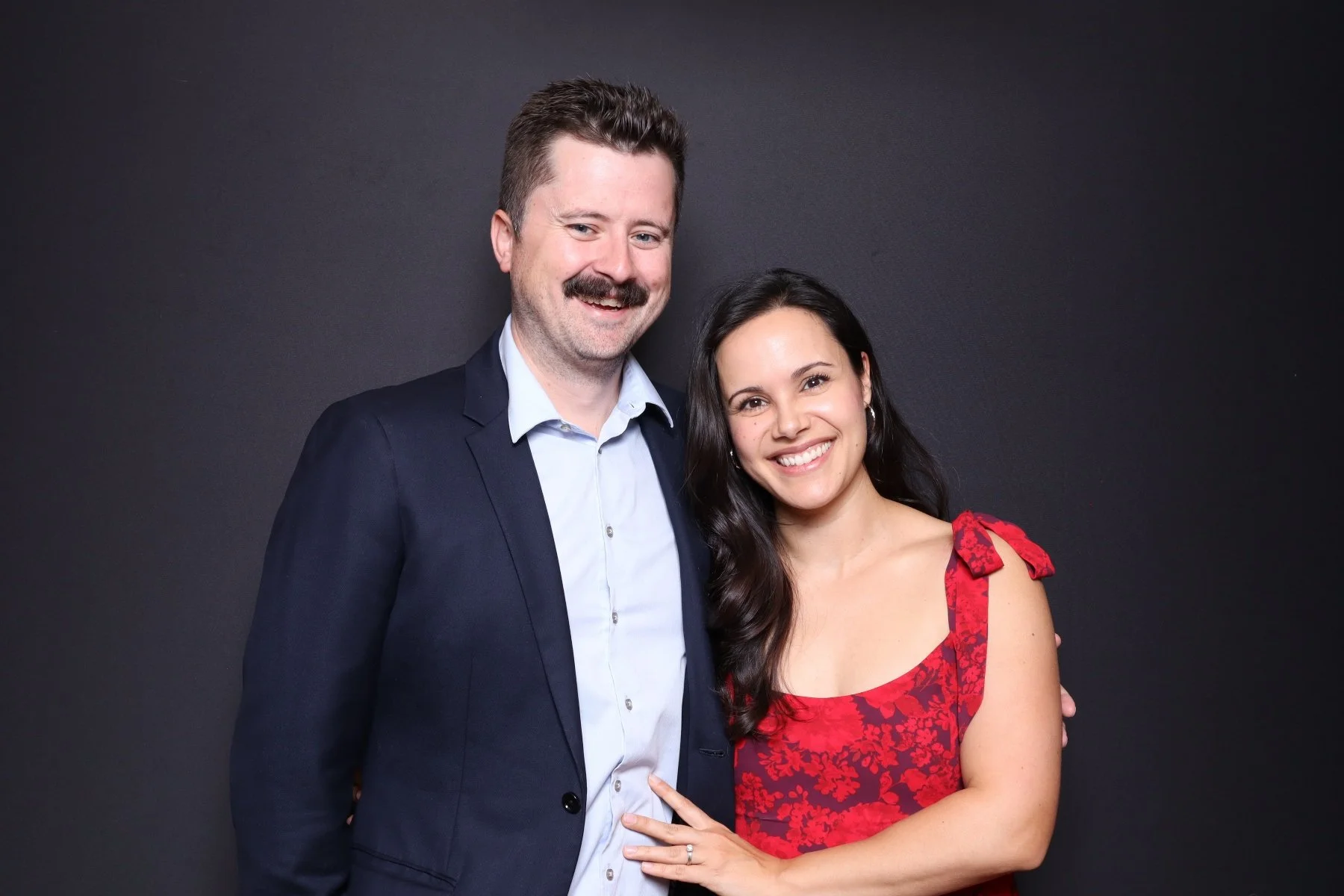 A smiling man and woman standing close together against a dark background, the man wearing a navy suit and blue shirt, and the woman in a red dress with floral pattern and shoulder bows.