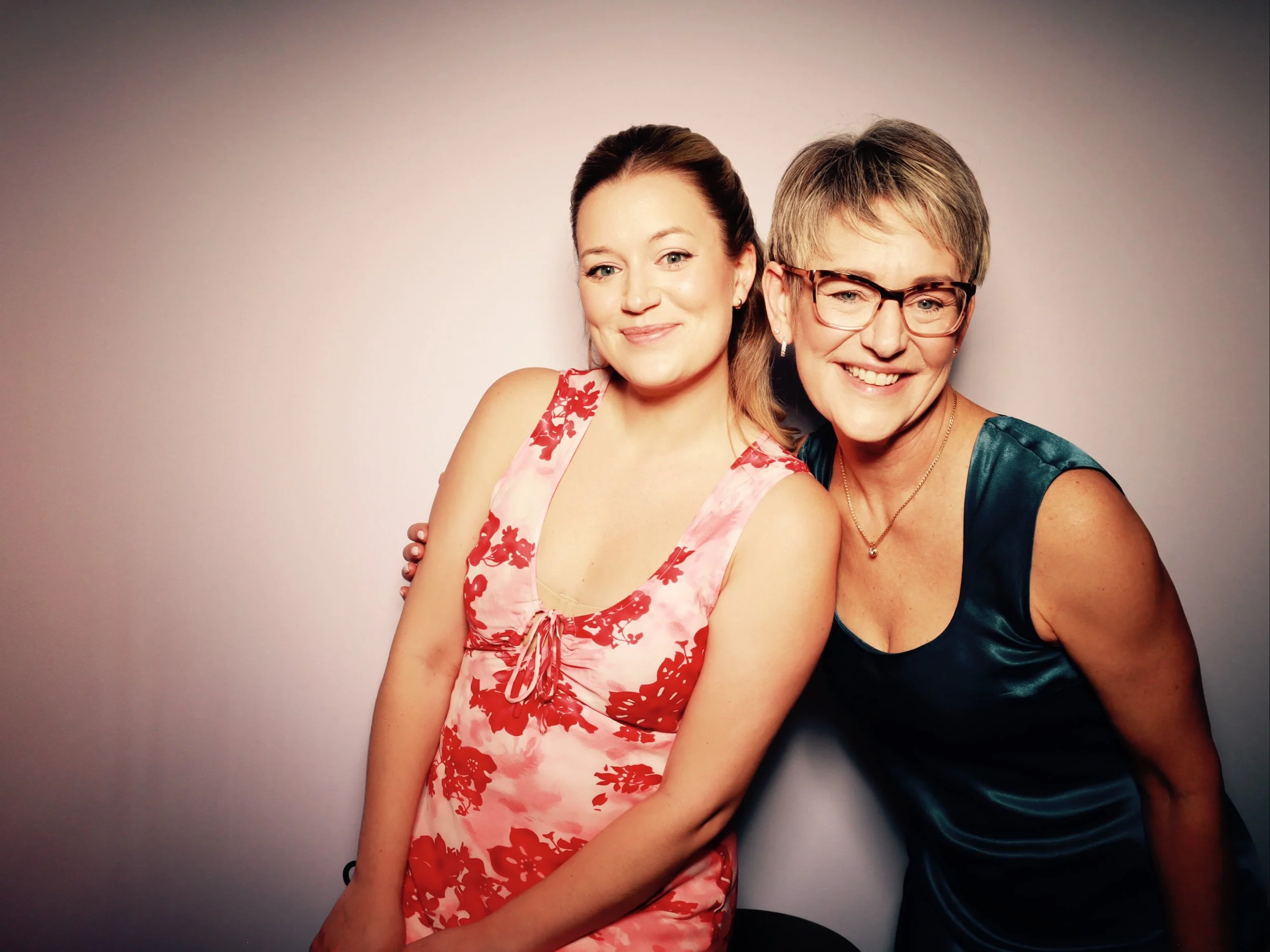 Two smiling women, one younger and one older, wearing dresses, posing together against a plain background.