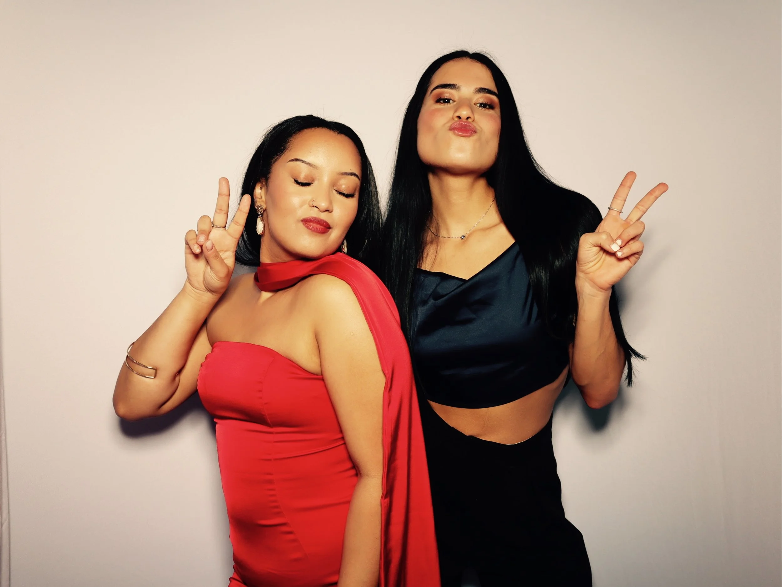 Two women posing together making peace signs, one in a red dress with earrings and jewelry, the other in a black satin crop top and skirt, both with long dark hair and confident expressions.