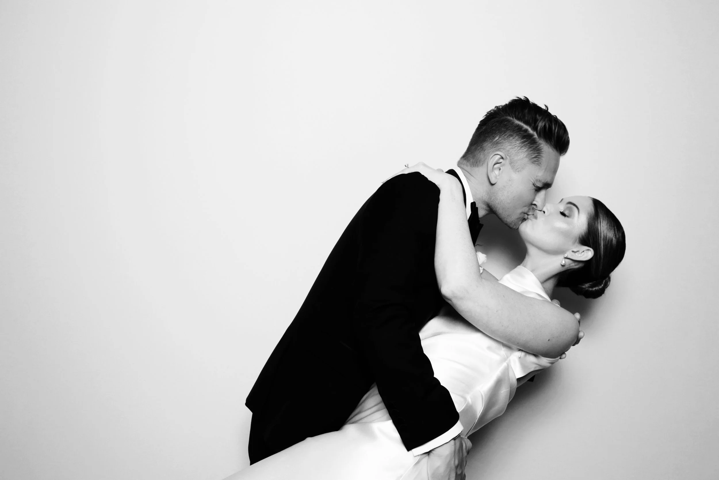 Black and white photo of a couple kissing, with the groom holding the bride in his arms against a plain background.