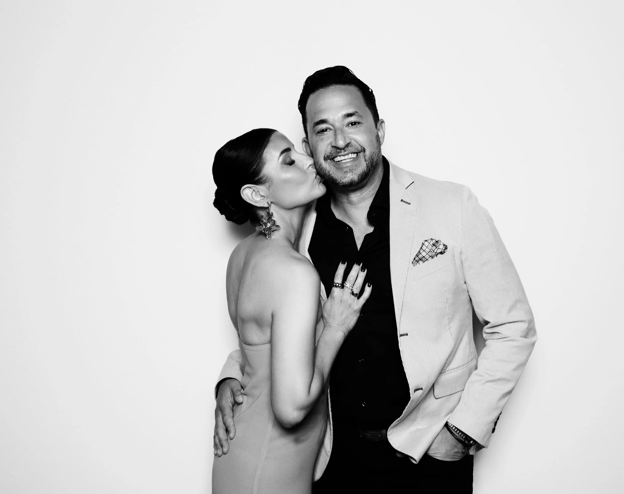 A woman kisses a smiling man on the cheek, both dressed formally, against a plain white background.