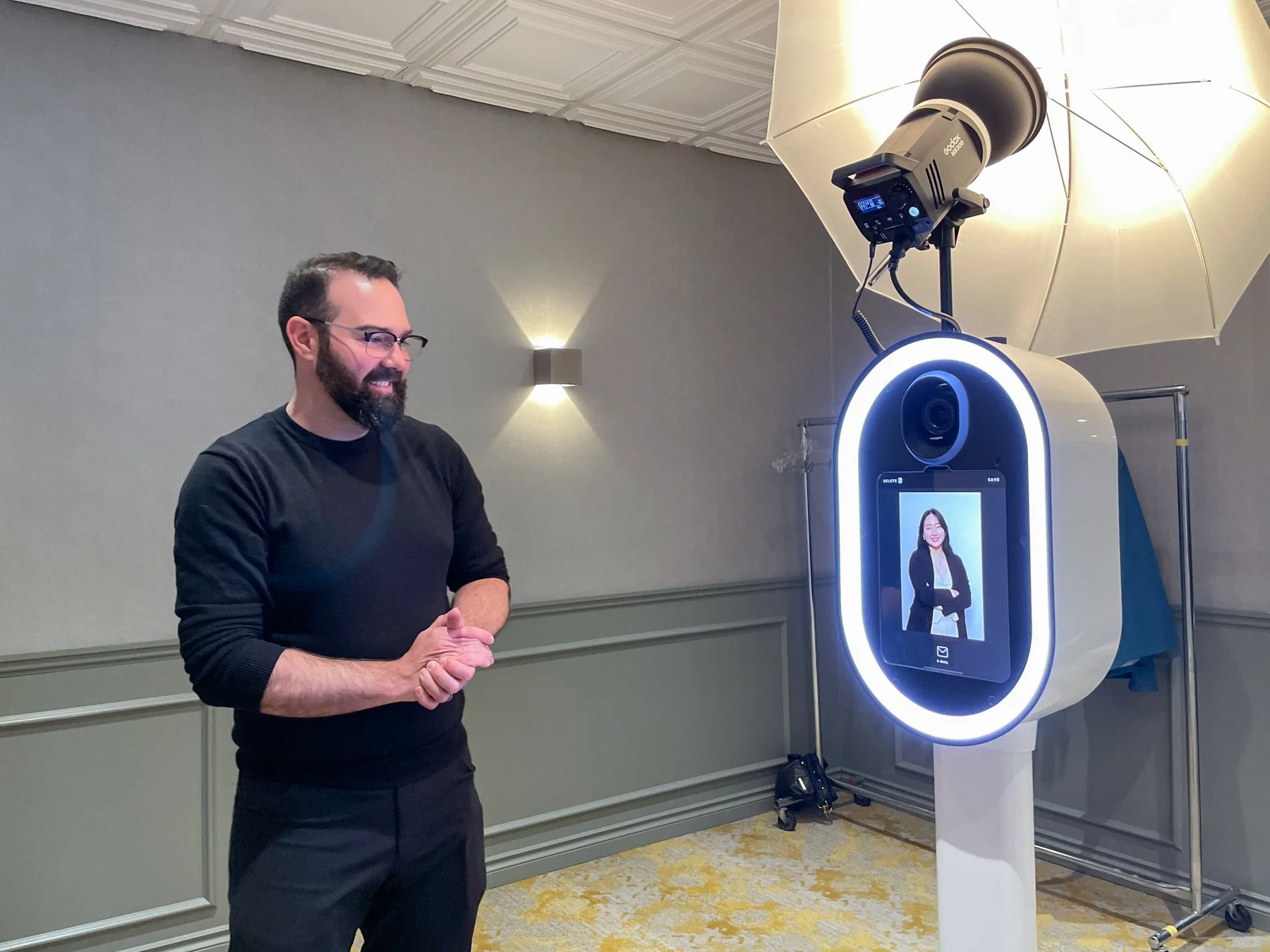 A man with glasses and a beard smiling and looking at a photo booth that has a digital screen displaying a woman in a blazer with arms crossed. The photo booth has a ring light, a camera, and a large umbrella light.