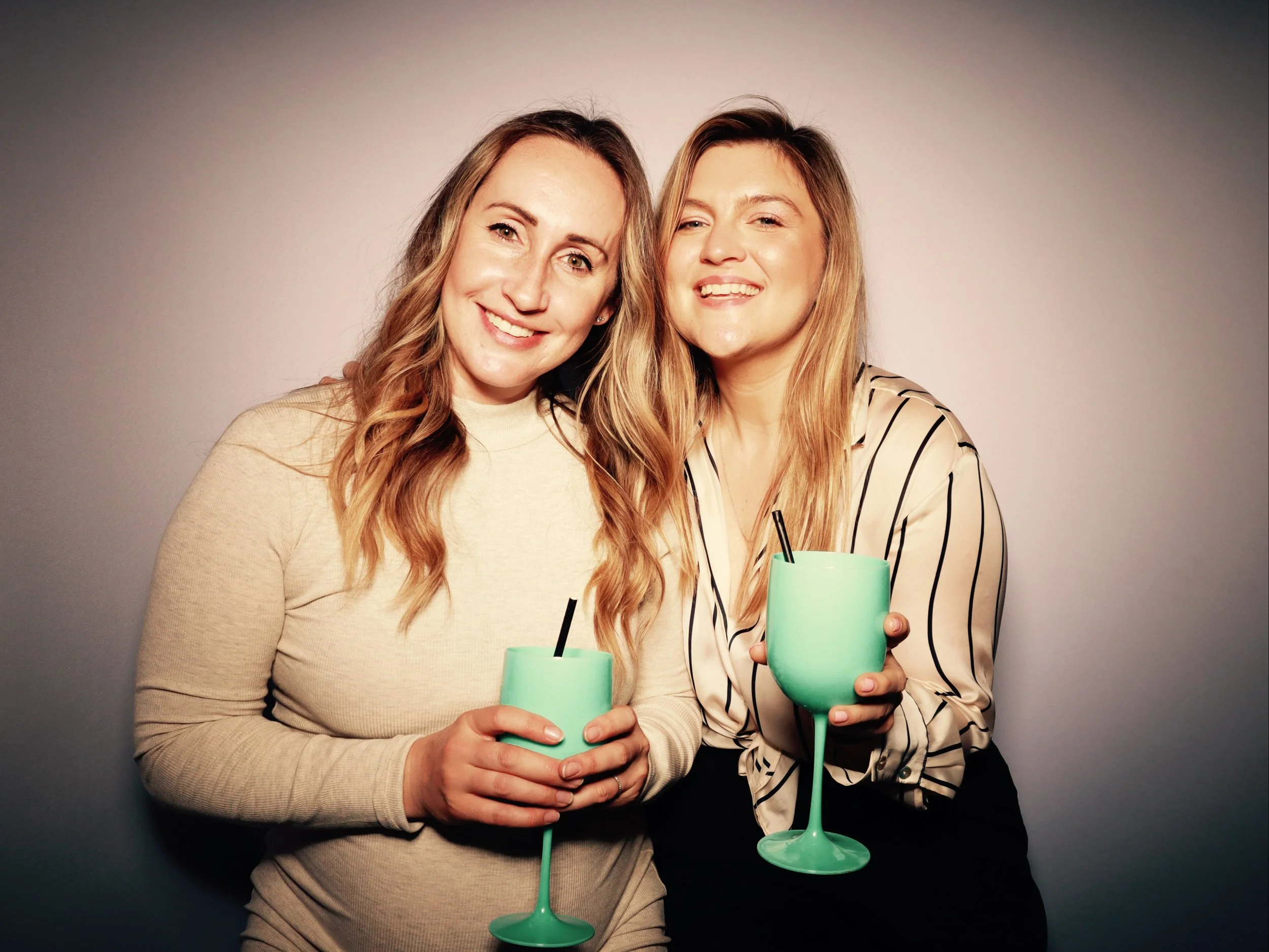 Two women smiling and holding turquoise wine glasses with black straws, standing close together against a neutral background.