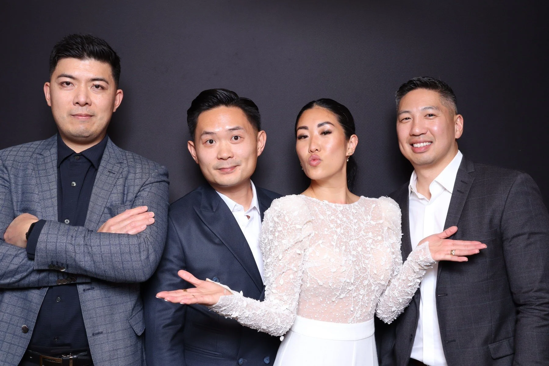 Four stylishly dressed people posing against a dark background, with one woman in a white lace top and three men in suits.