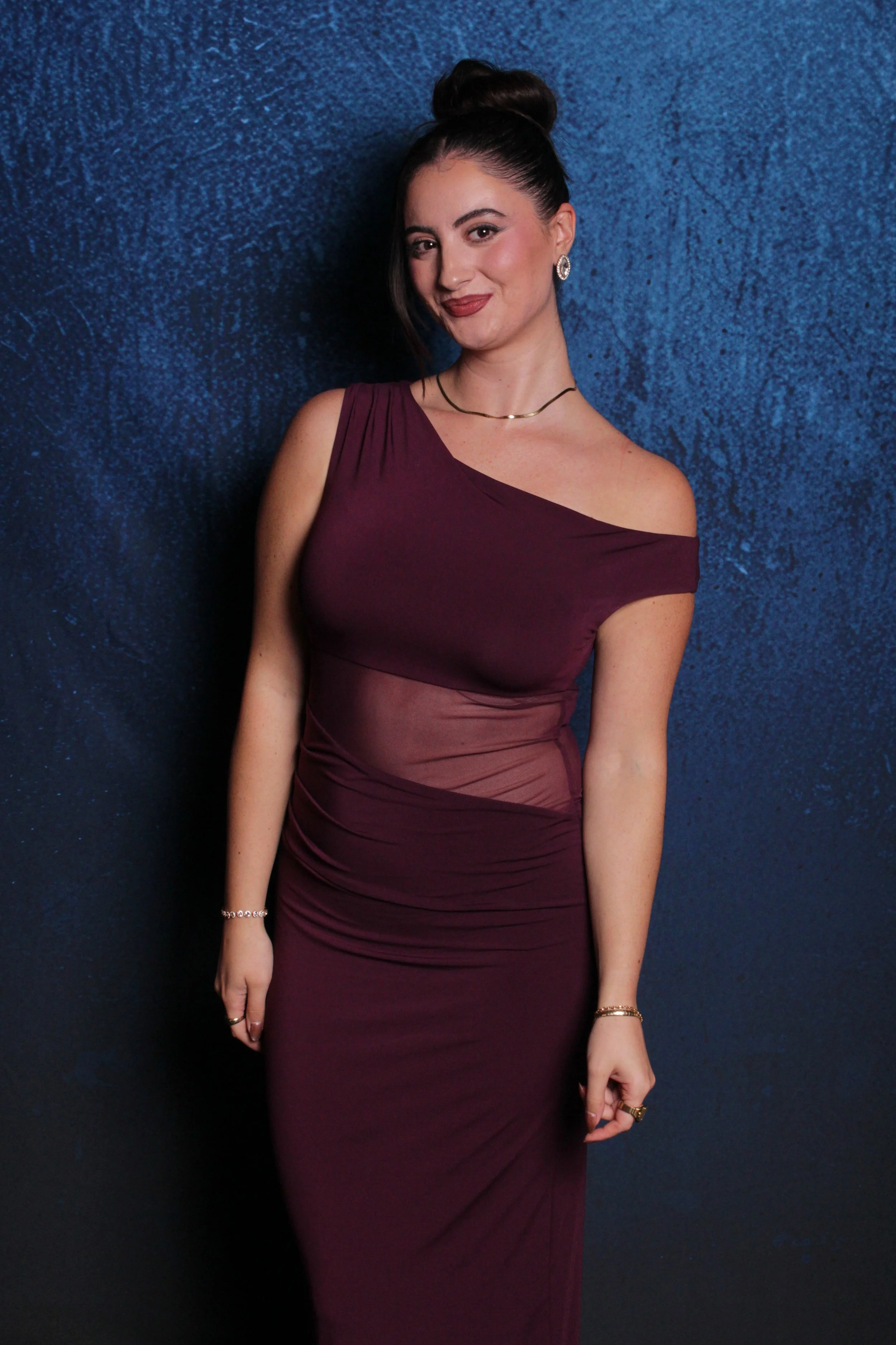 A young woman with dark hair styled in an updo, wearing a burgundy, off-shoulder, form-fitting dress with sheer panels, standing against a textured dark blue background.