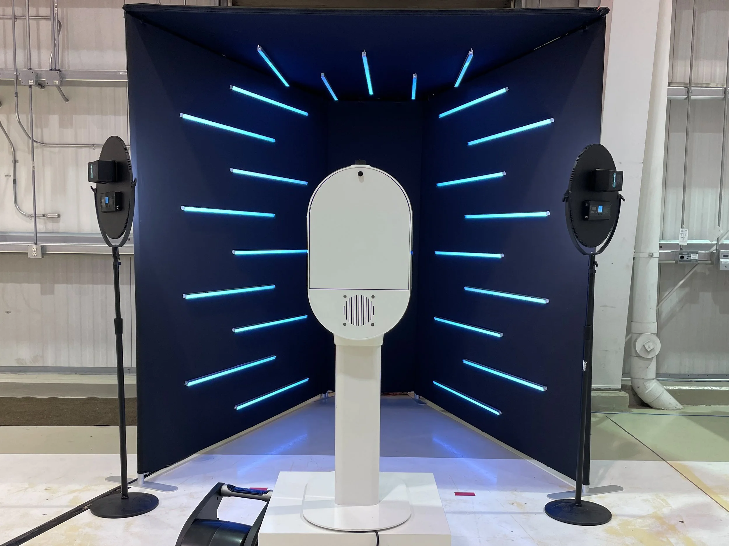A white virtual reality headset on a pedestal in front of a dark blue wall with blue LED lights, flanked by two black speakers on stands.
