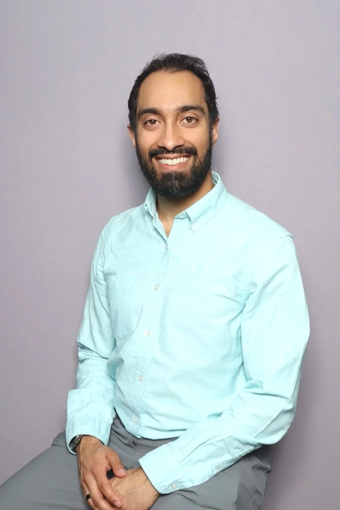 A man with dark hair, beard, and light skin, wearing a light blue button-down shirt and gray pants, smiling and sitting against a plain light gray background.