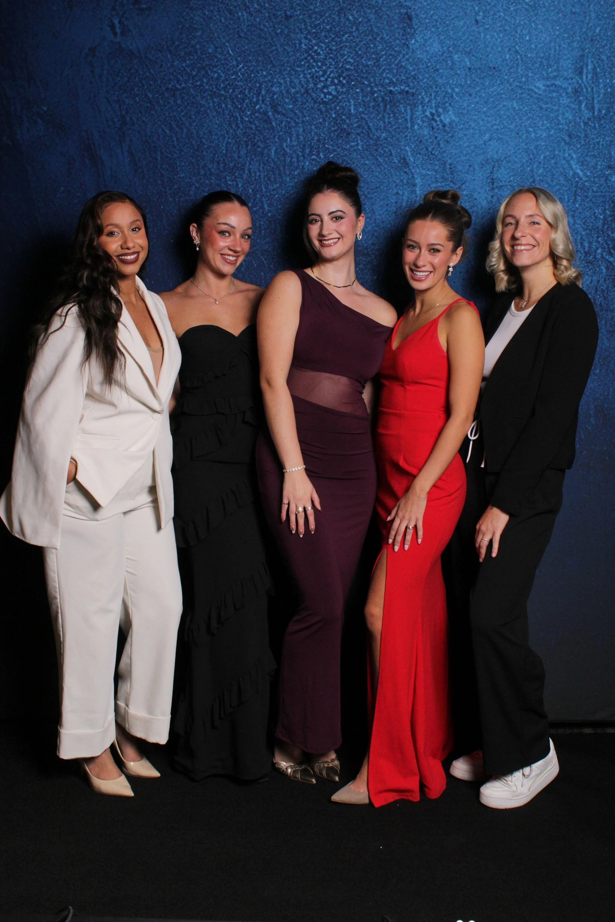 Five women in formal dresses posing together against a dark blue textured background at a social event.