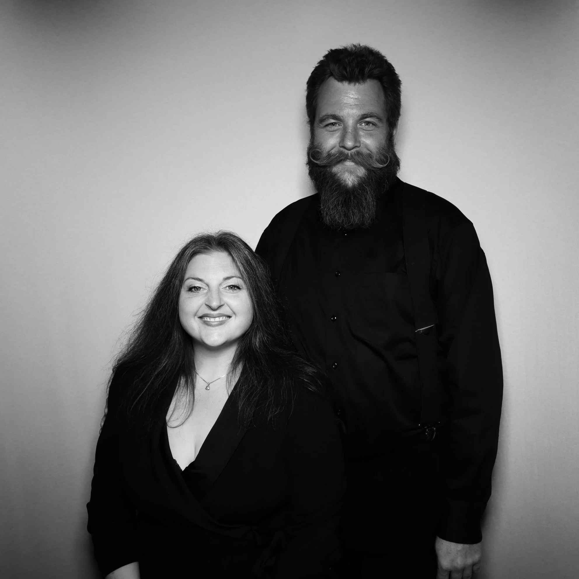Black and white photo of a smiling woman with long hair and a man with a beard and mustache, both dressed in black, standing against a plain background.