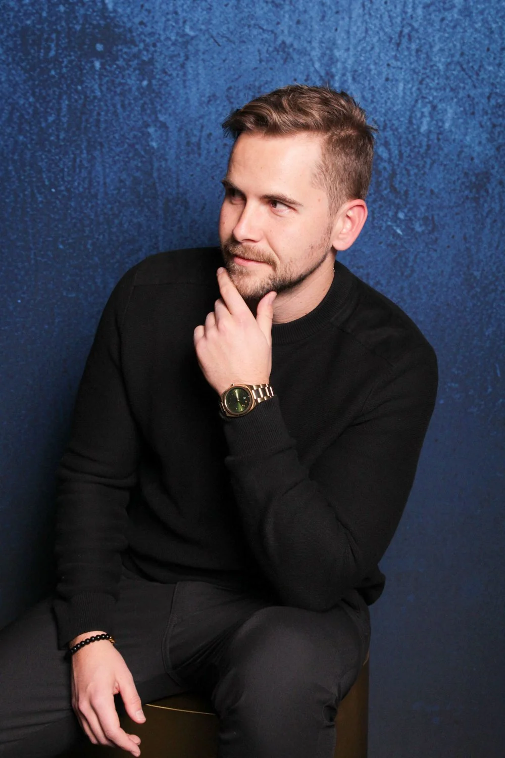 A man sitting against a blue textured wall, wearing a black sweater, a wristwatch, and a beaded bracelet, with his hand resting on his chin.