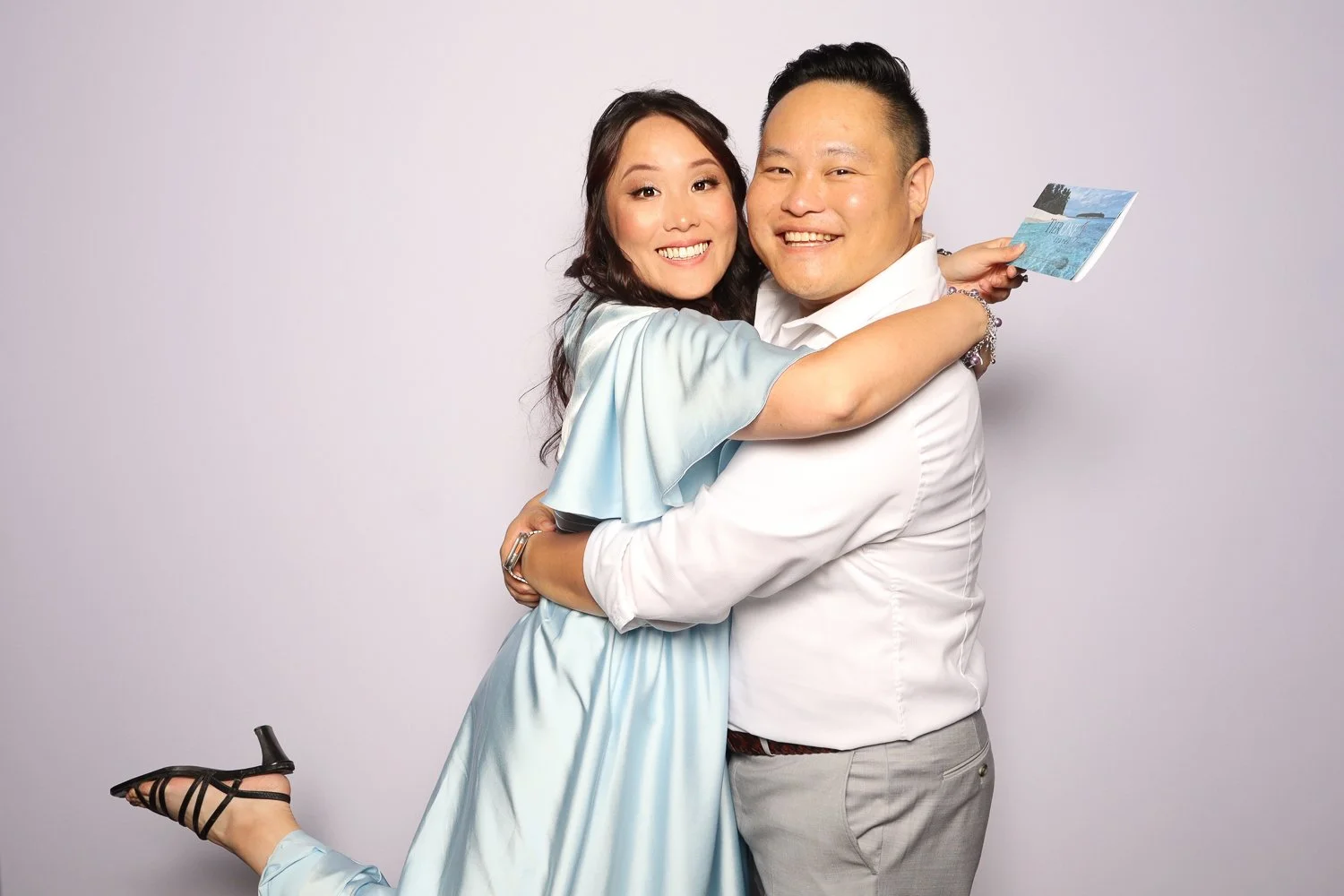 A happily smiling couple hugging, the woman with long dark hair wearing a light blue dress and black heels, and the man with short dark hair in a white shirt and light gray pants, standing against a plain white background.
