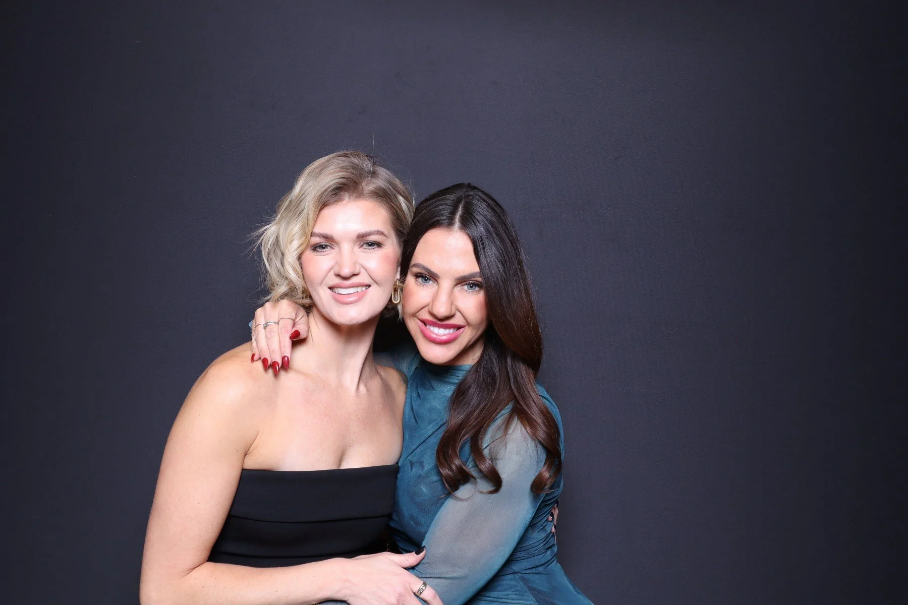 Two women hugging and smiling at the camera against a dark background.