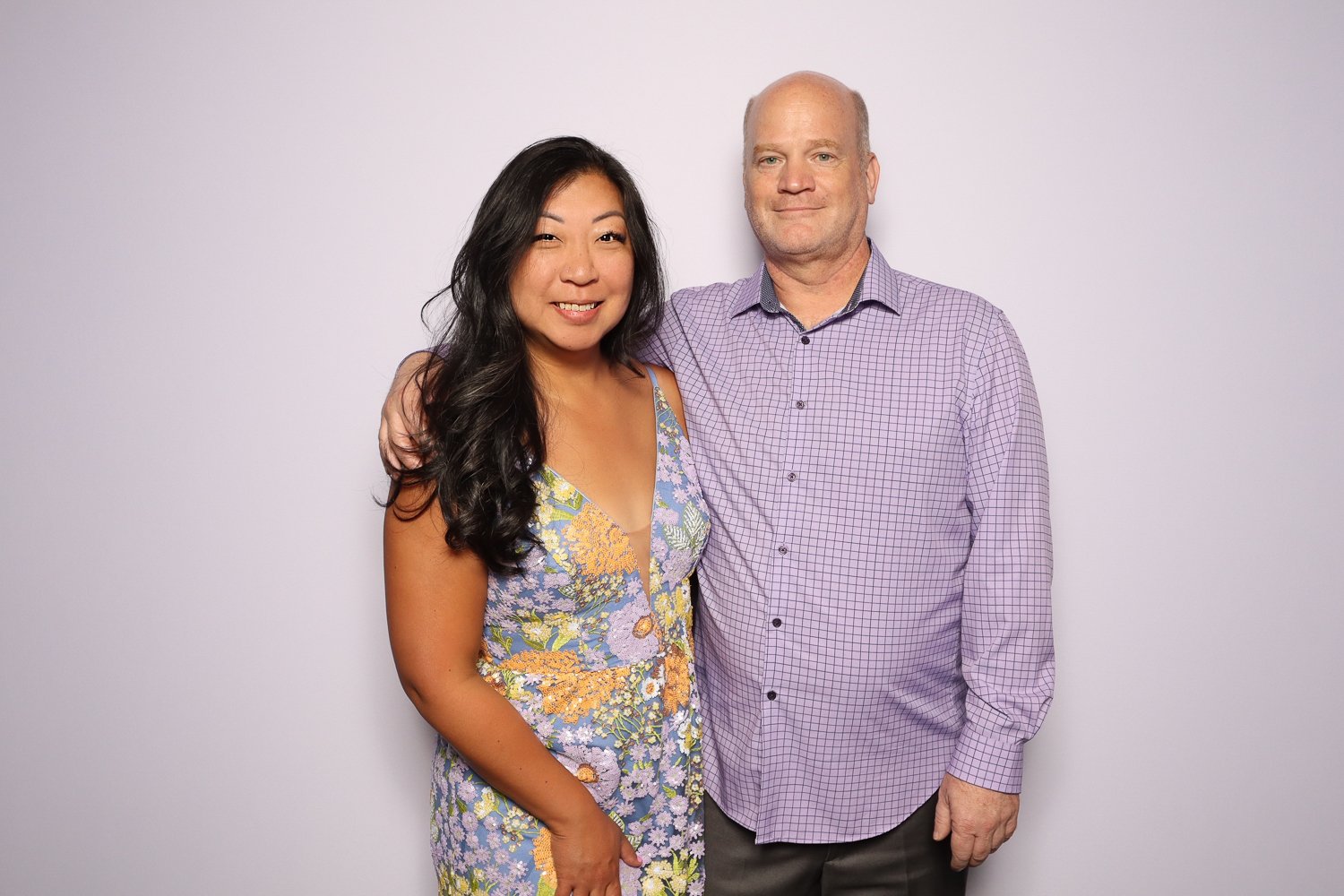 A woman with long black hair wearing a colorful floral dress standing next to a man with short blond hair and a purple checkered shirt against a light purple background.