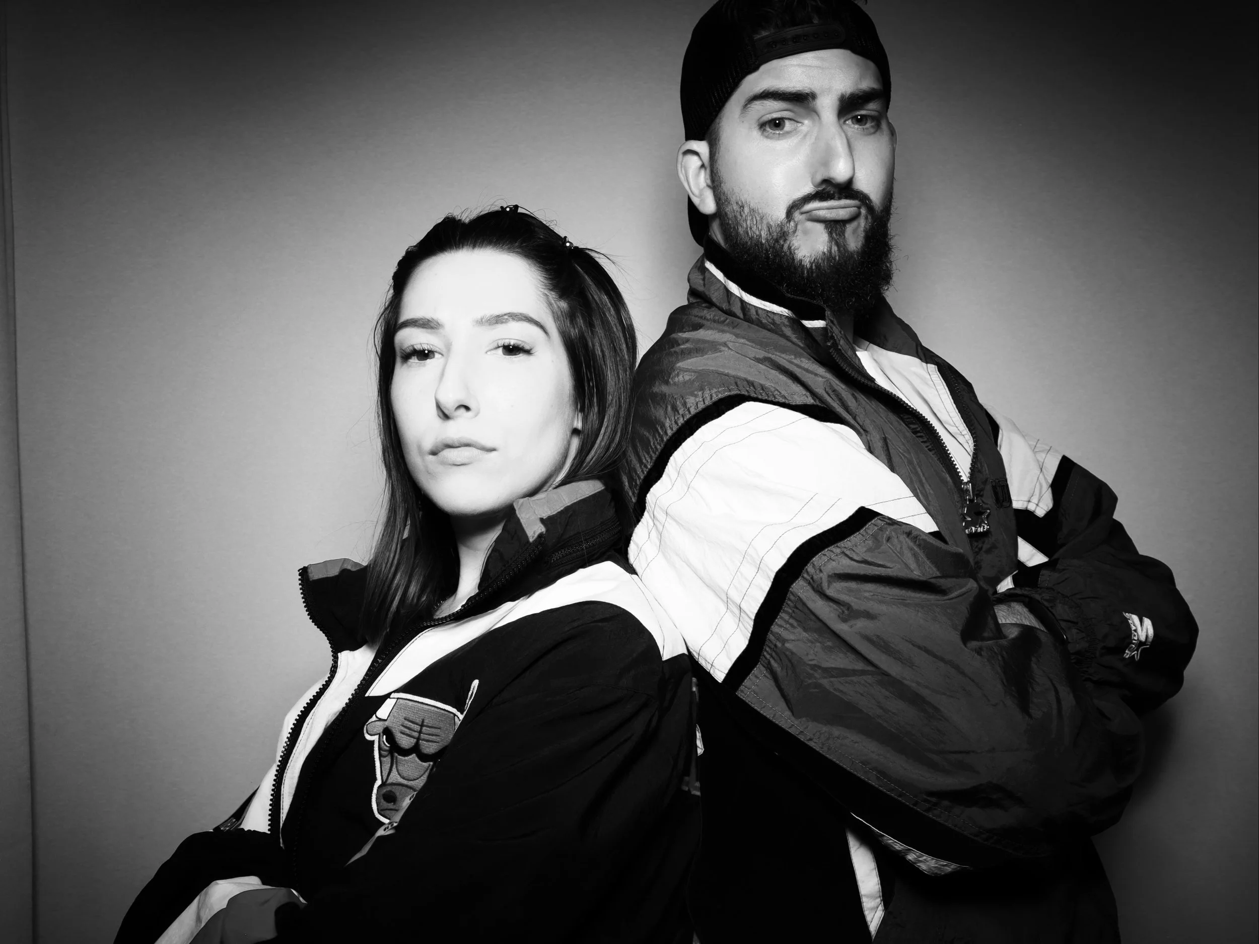 Black and white photo of a woman and man standing back-to-back with arms crossed, both wearing jackets. The woman has shoulder-length hair, and the man has a beard and is wearing a beanie.