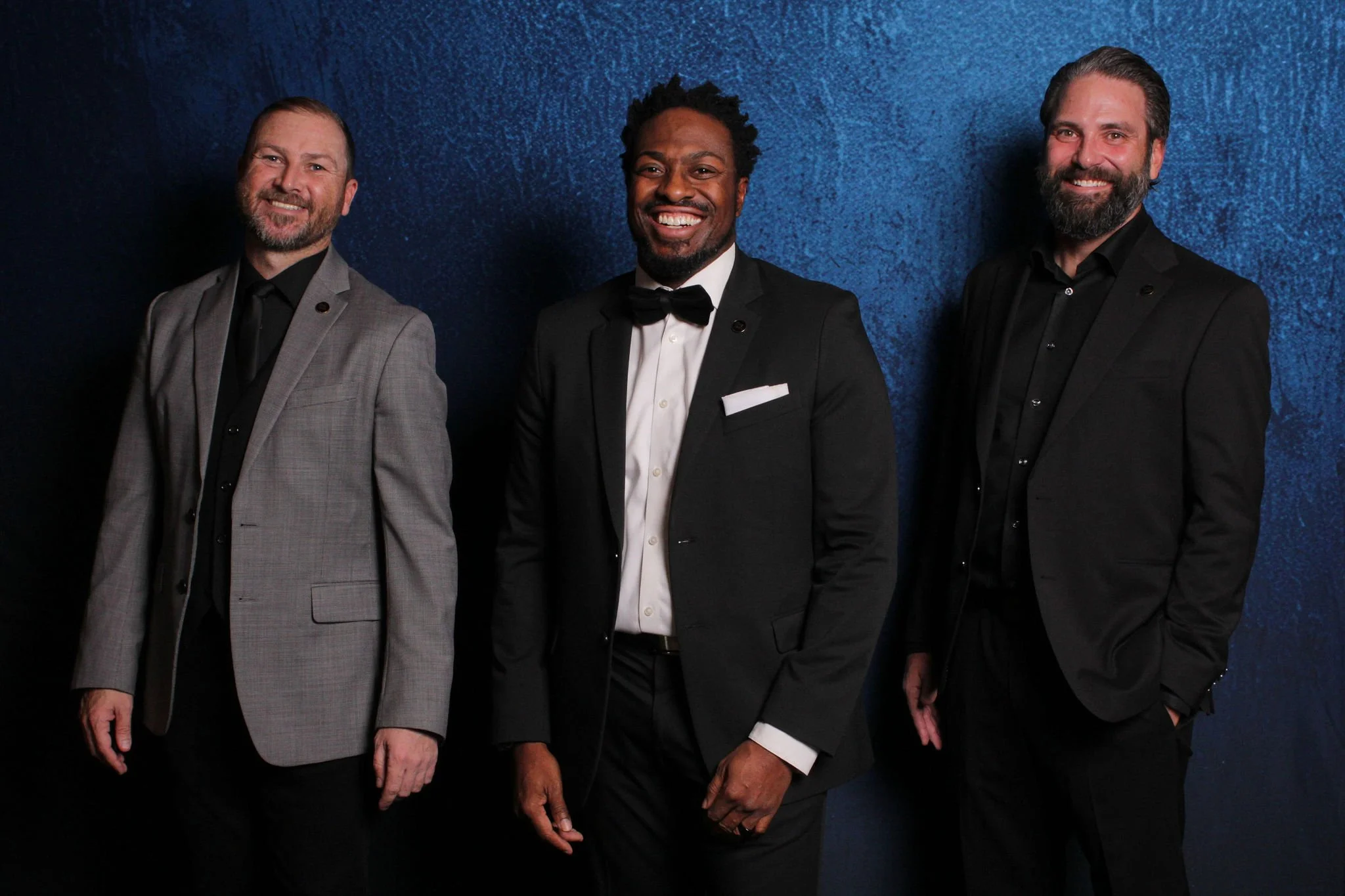 Three men dressed in formal black tuxedos with different blazer colors, standing against a dark blue textured background, smiling at the camera.