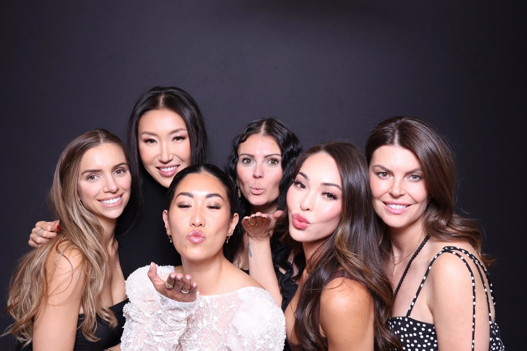 Group of seven women posing together in front of a black background, smiling and making kissing faces.