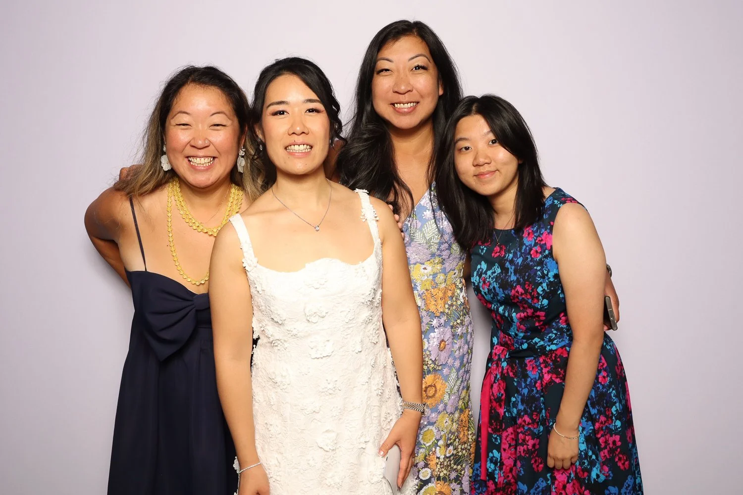 Four women smiling and posing together against a plain light background, dressed in colorful dresses.