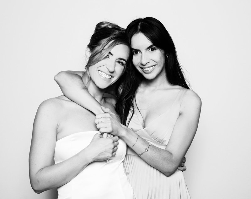 Two women embracing and smiling, one winking, against a plain background.