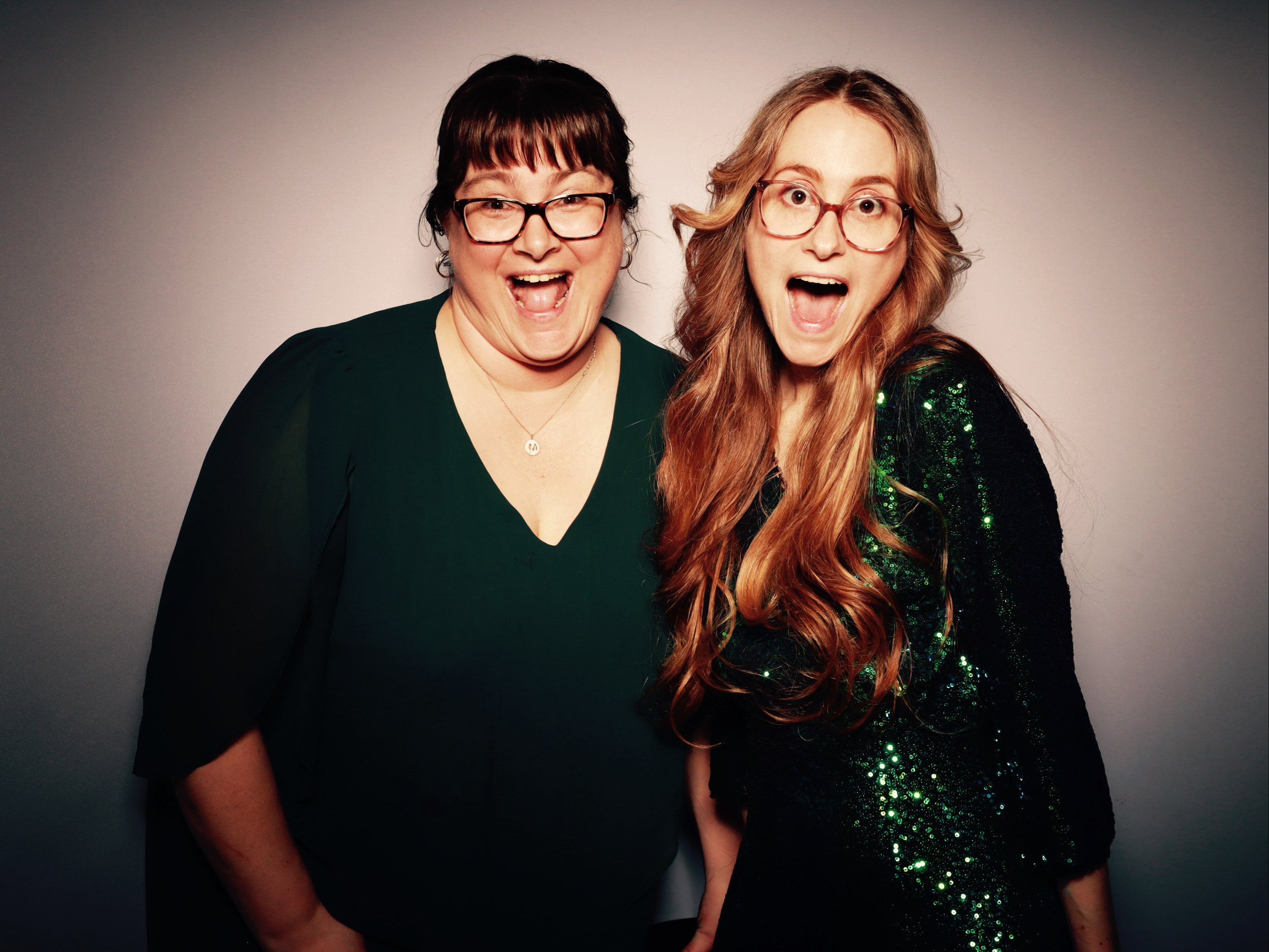 Two women with glasses smiling and making surprised expressions, standing close together against a neutral background.