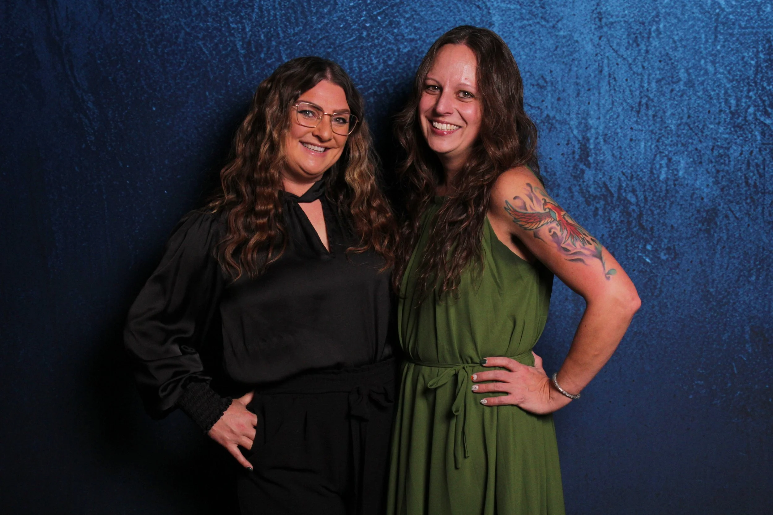 Two women standing together against a blue textured background, smiling at the camera. One woman has long, wavy brown hair, glasses, and is wearing a black blouse. The other woman has long, wavy hair, a colorful tattoo on her right arm, and is wearin