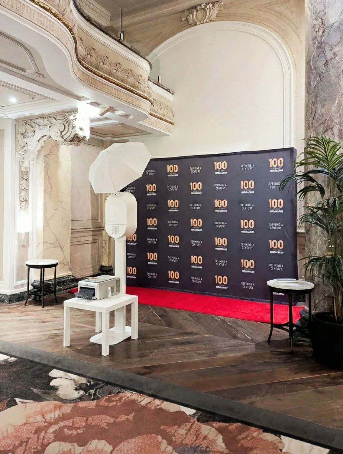Event backdrop with logos, photo booth equipment including reflector and projector, small tables, potted plant, ornate venue interior with marble and wood flooring, and a red carpet.