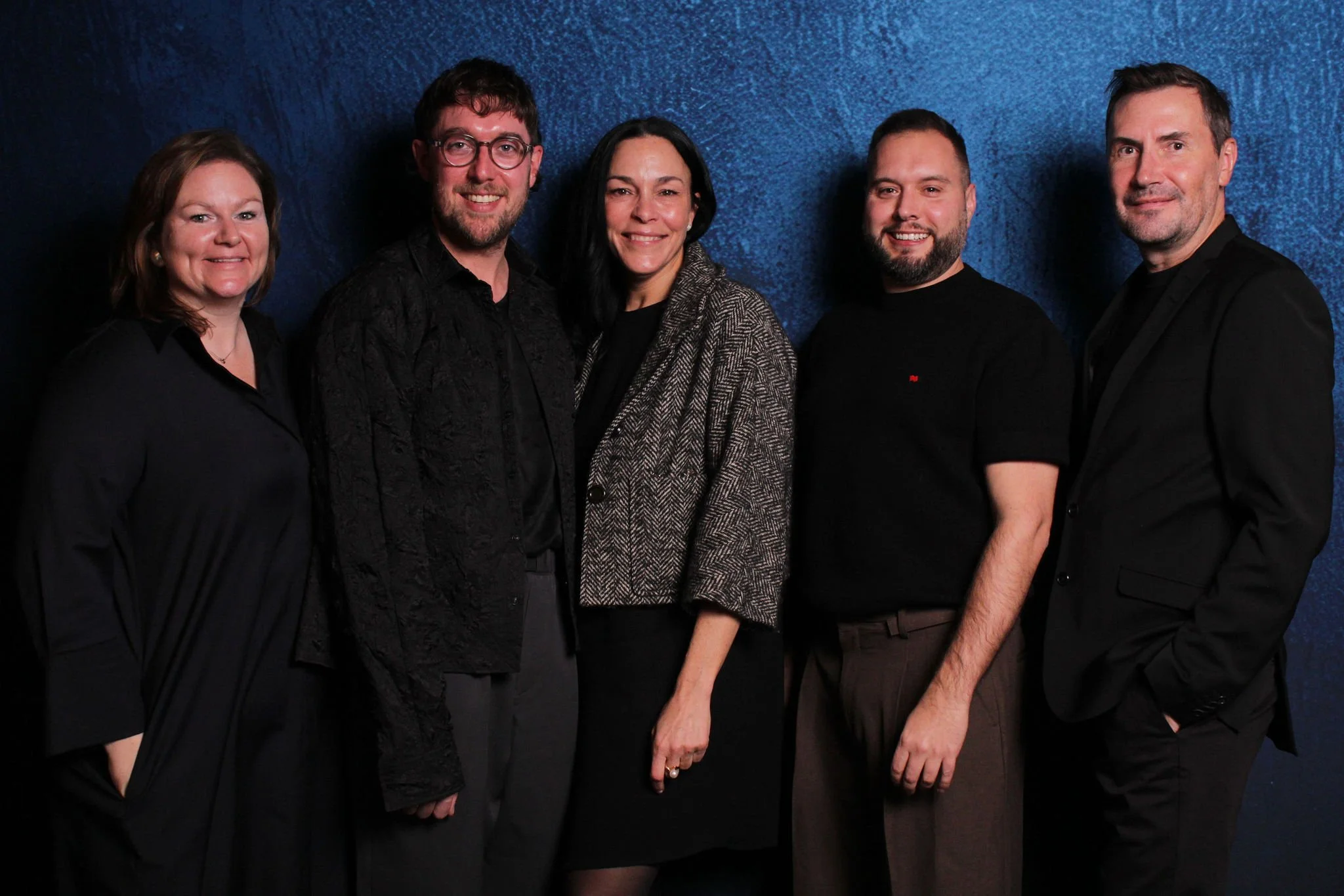 Group of five people standing together in front of a blue textured background, smiling at the camera.