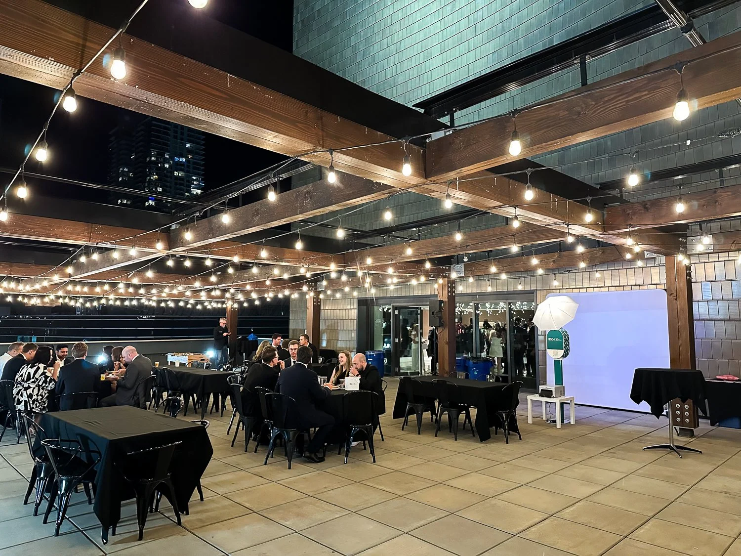 Nighttime gathering of people on a rooftop patio with string lights, tables, chairs, and photography equipment.