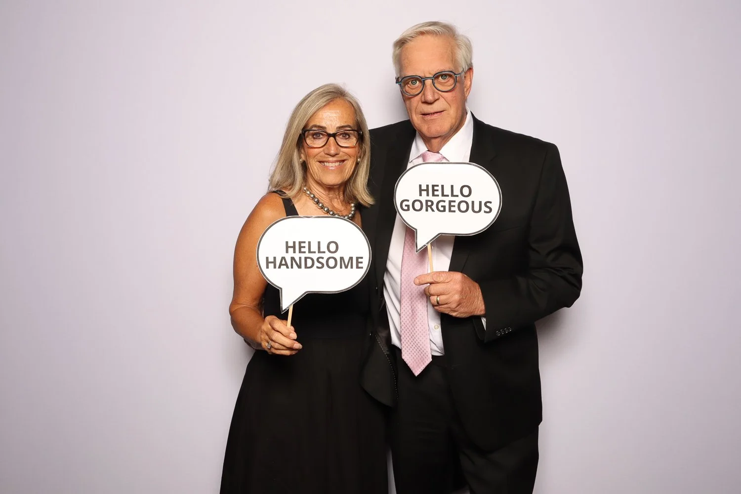 An elderly couple dressed in formal attire standing against a light background, holding speech signs that read "Hello Handsome" and "Hello Gorgeous".