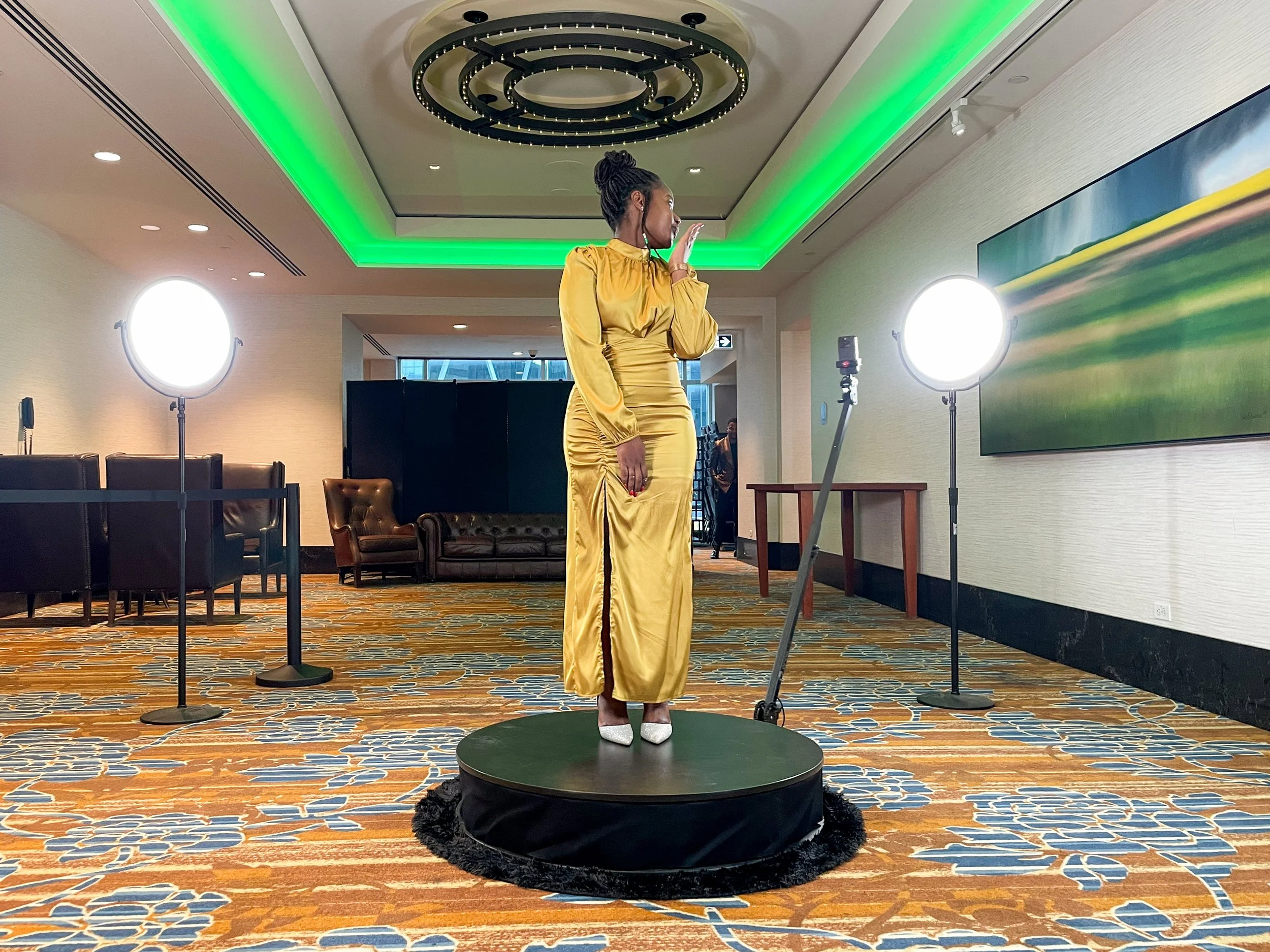 A woman in a gold satin dress and white shoes standing on a small circular platform, with her hand near her face, in a well-lit indoor space with modern decor and green lighting accents.