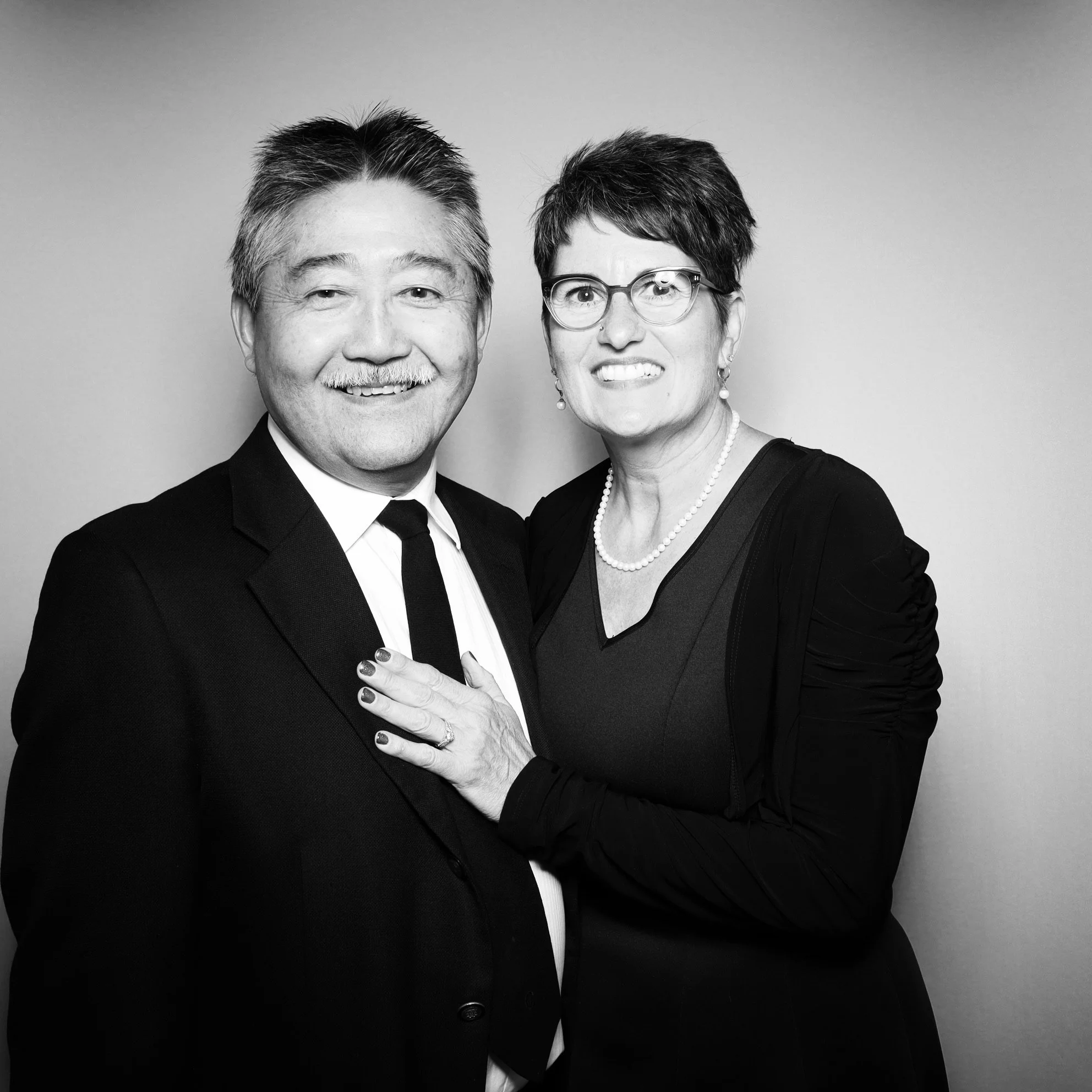 Black and white photo of a smiling man in a suit and a woman with short hair, glasses, and a pearl necklace, standing close together.
