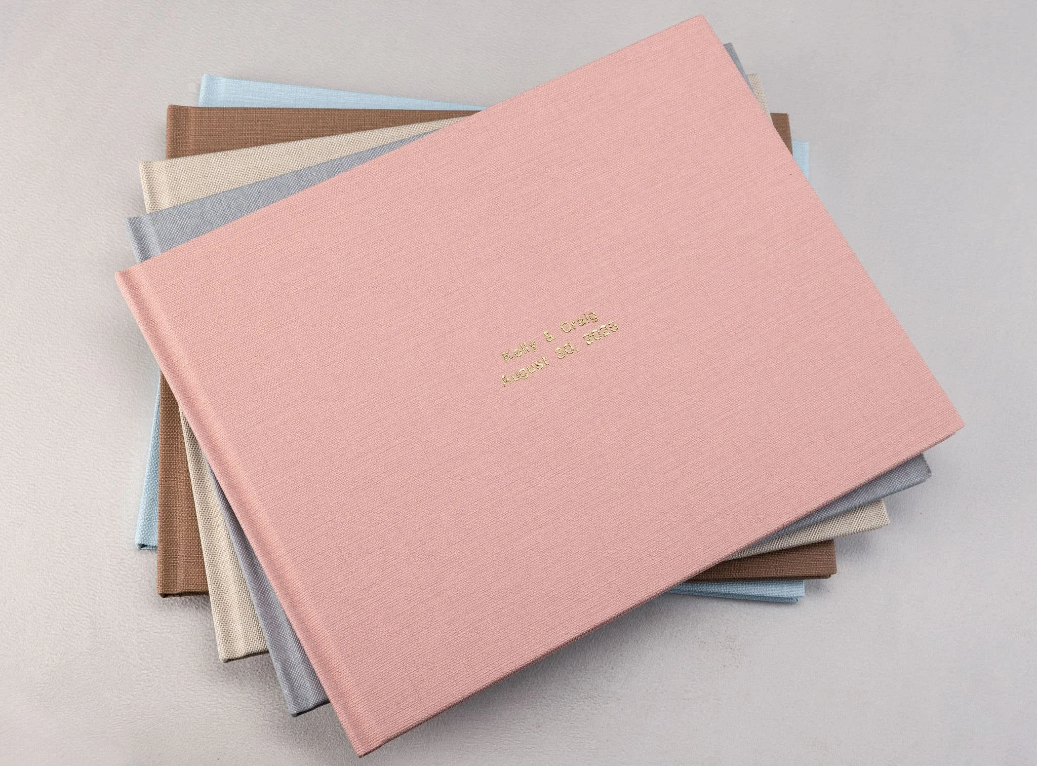 Stack of 6 wedding photo albums with pastel-colored covers, featuring a pink album on top with gold embossed text, placed on a light surface.