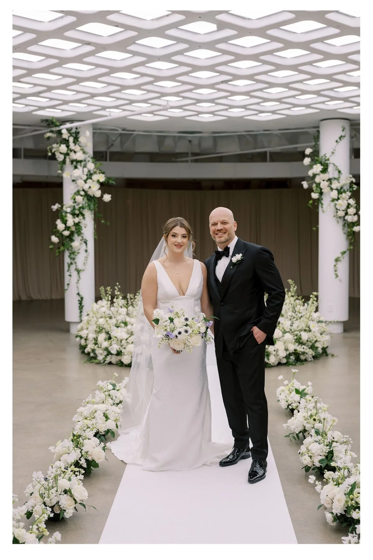 A bride and groom standing on a white aisle runner decorated with white flowers, at a wedding ceremony. The bride is in a white gown holding a bouquet, and the groom in a black tuxedo with a bow tie. Behind them are white floral arrangements and floral decorations on white pillars, in a modern indoor venue.
