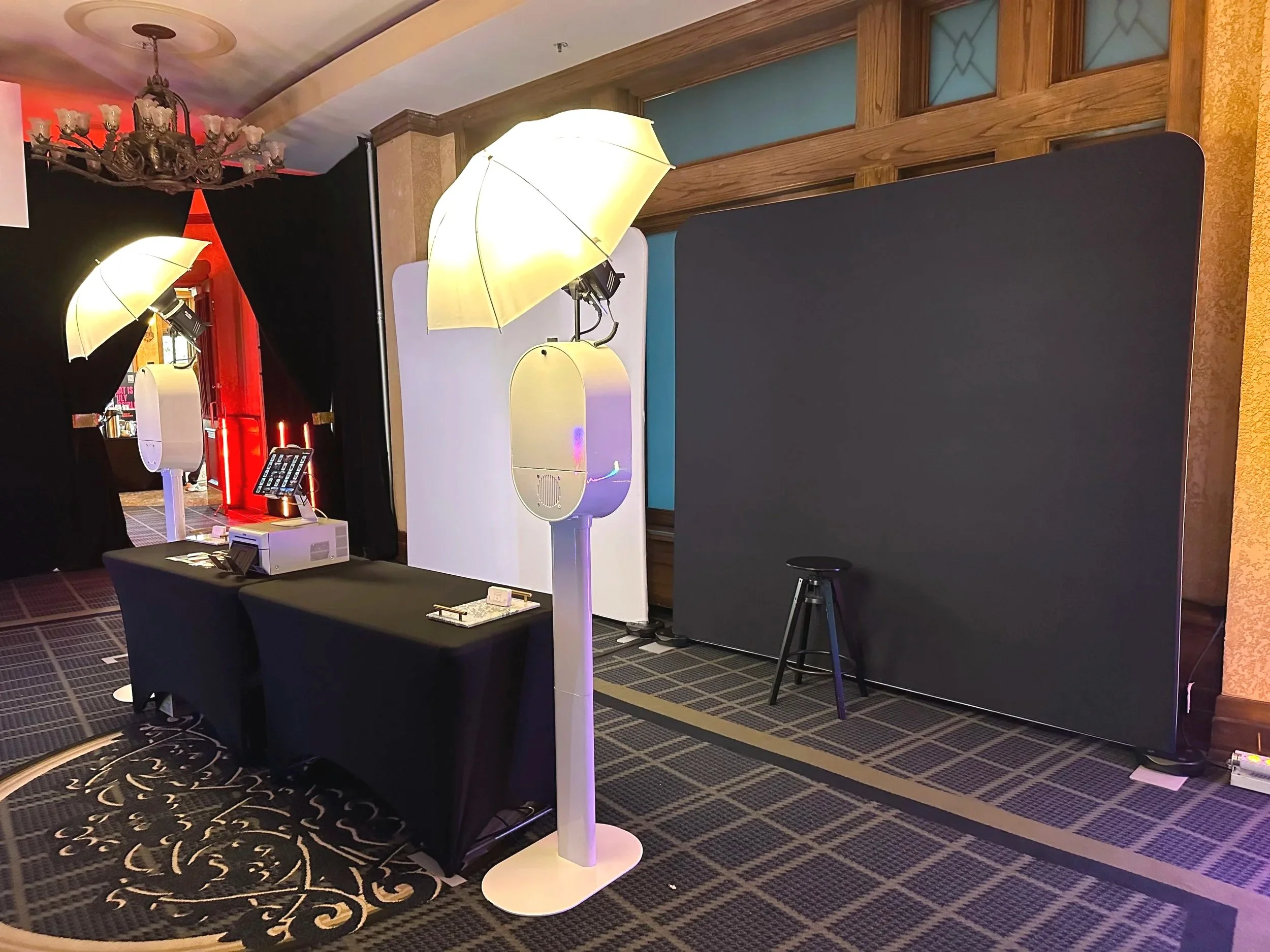 An indoor space with black, white, and gold decor, including two unique floor lamps with umbrellas, a black stool, and a black table with items on it. There is a round, patterned carpet in the foreground and a black backdrop with a chandelier hanging from the ceiling.