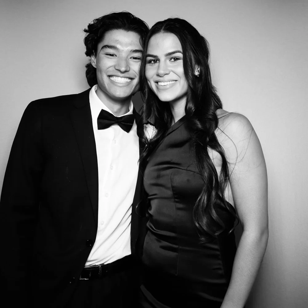 A smiling young man in a tuxedo with a bow tie and a young woman in a satin dress, standing close together in black and white photo.