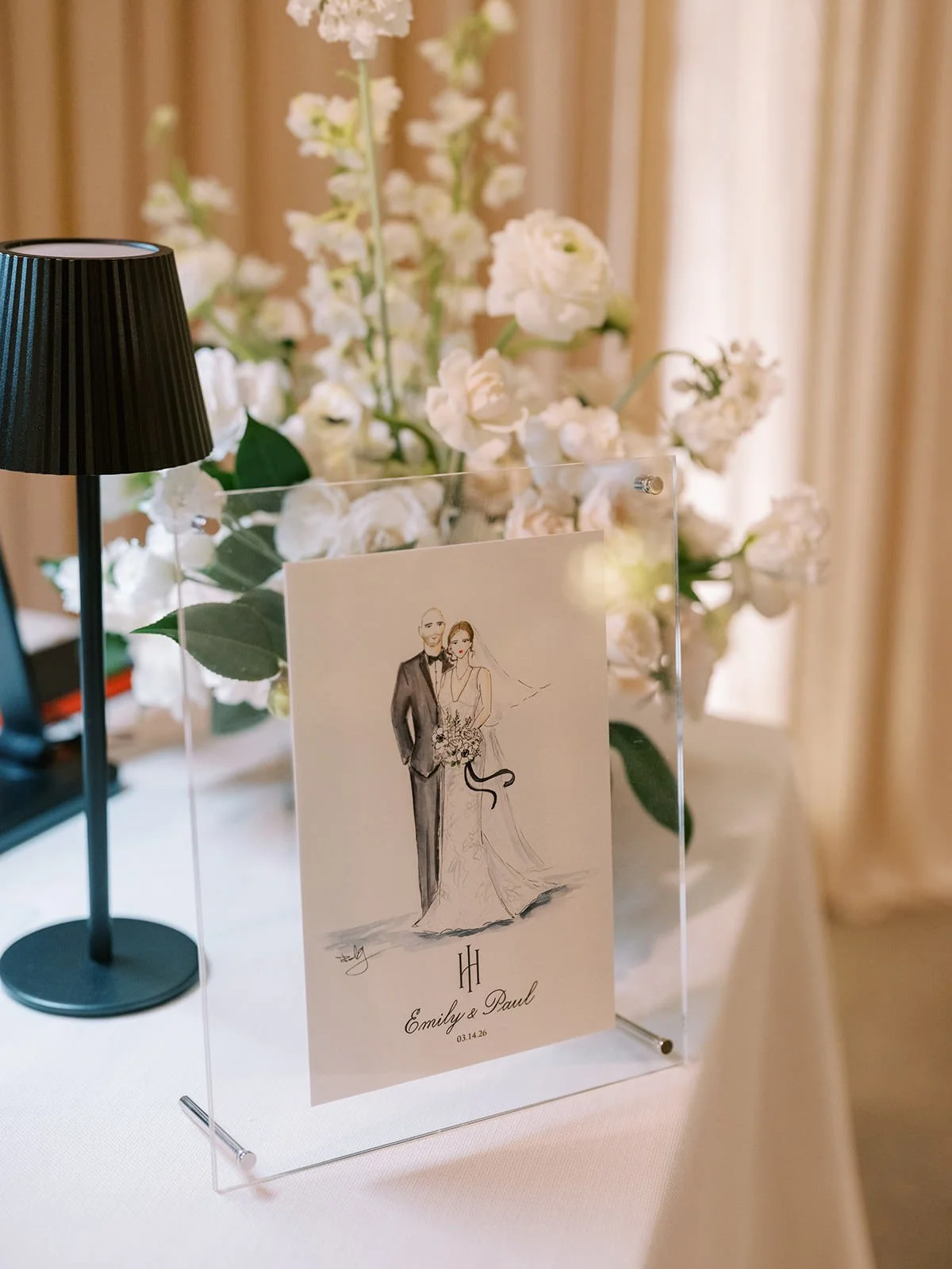 Wedding invitation card with a sketch of a bride and groom, surrounded by white flowers and a black table lamp in the background.