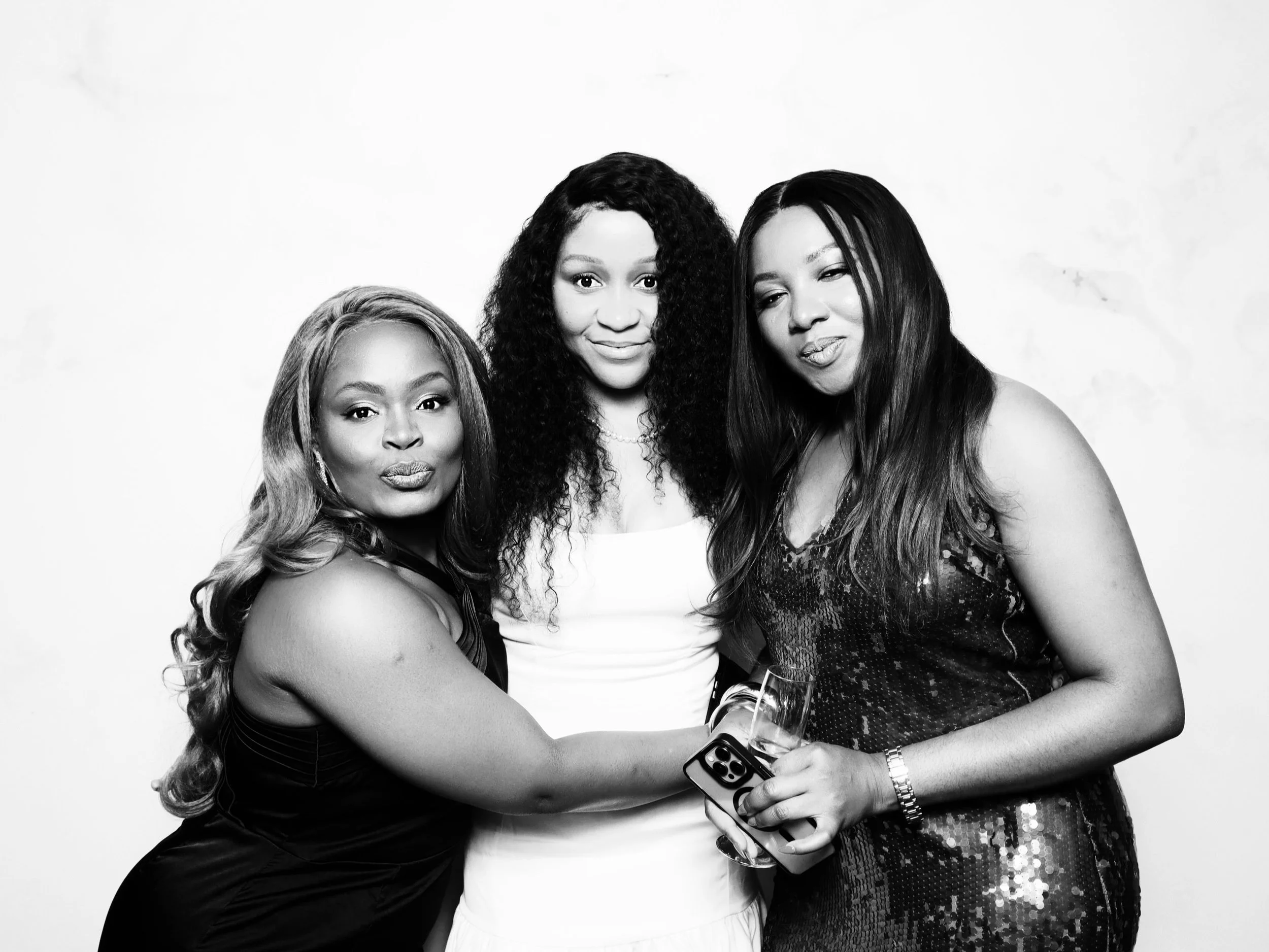 Three women of African descent dressed up, standing close together, with one holding a glass of champagne and a phone, posing for a black and white photo against a plain background.