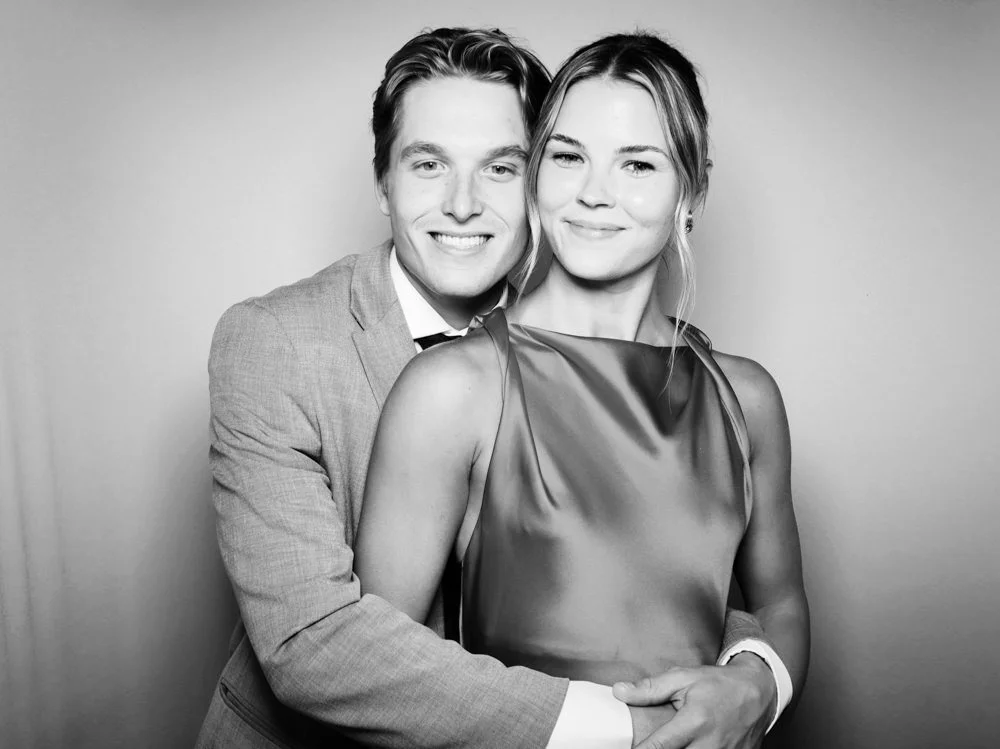 A black and white photo of a smiling man and woman standing close together, with the man hugging the woman from behind. The man wears a suit and tie, and the woman wears a satin sleeveless dress, both looking toward the camera.