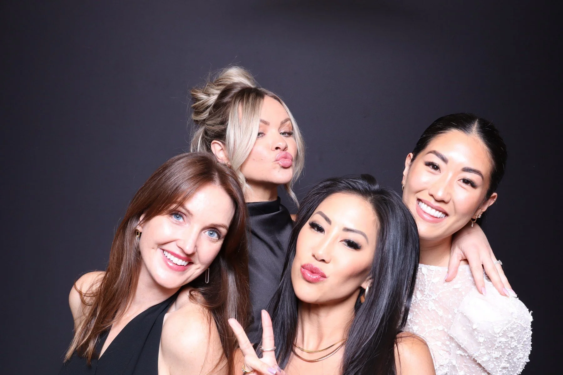 Four women posing together for a photo, smiling and making kissy faces, with a dark background.