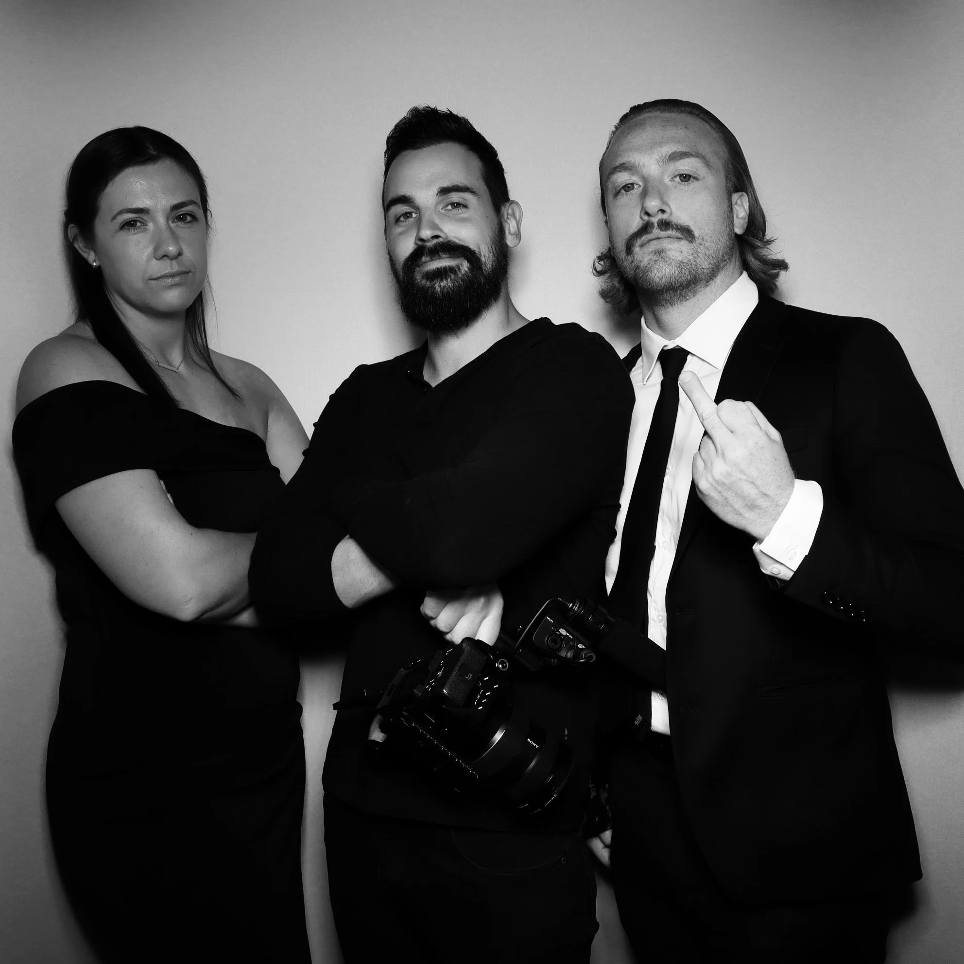 Three people posing against a plain wall, with a woman on the left, a man with a beard in the center, and a man in a suit on the right, with cameras hanging from his neck, the man on the right pointing his middle finger.