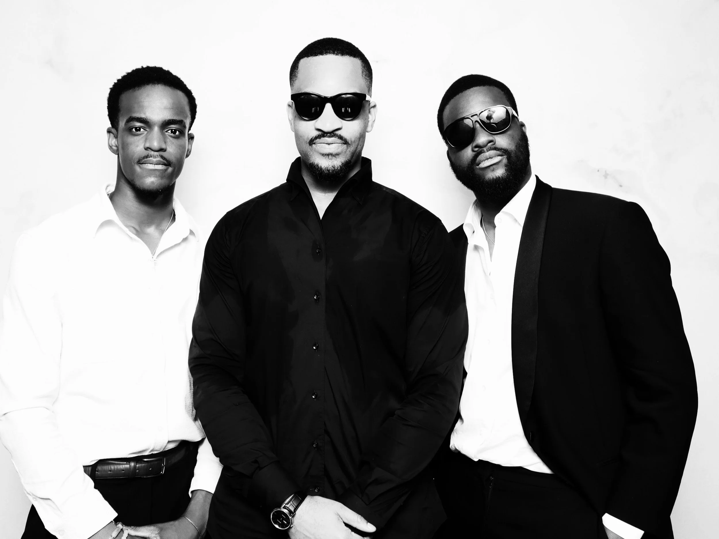 Three men dressed in formal attire, two wearing sunglasses, posing against a plain background in a black-and-white photograph.