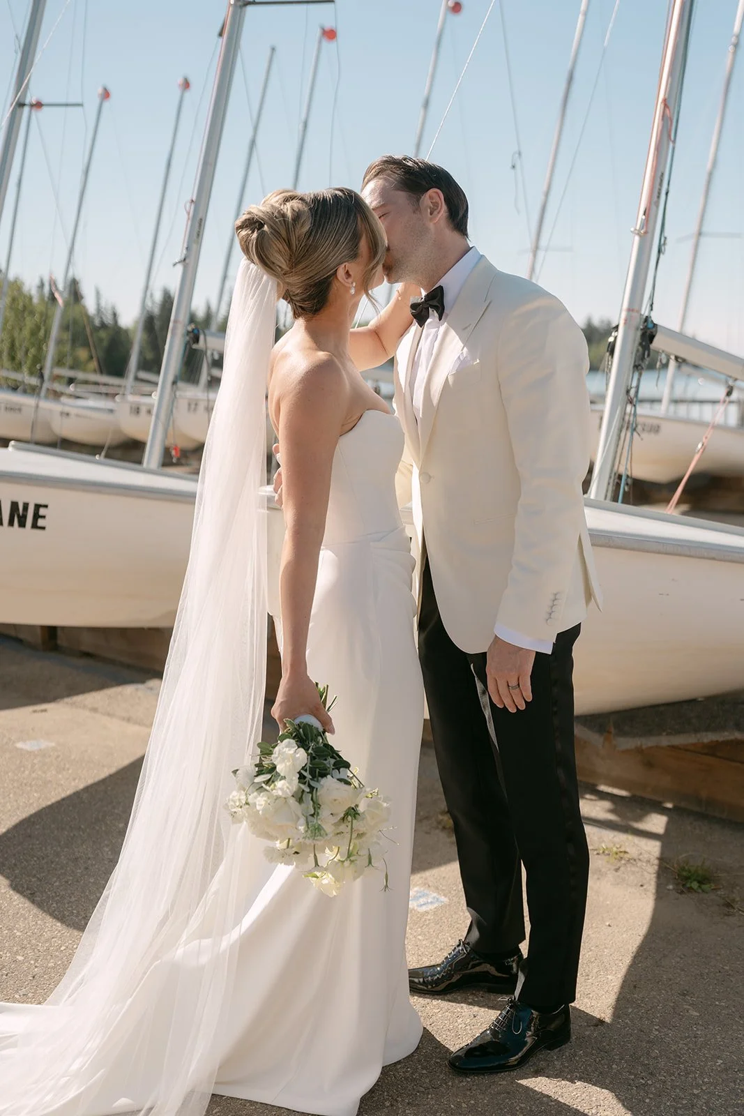 A bride and groom kiss at a marina with sailboats in the background. The bride wears a white strapless wedding gown and holds a bouquet of white flowers. The groom wears a white suit jacket, black pants, and a black bow tie.