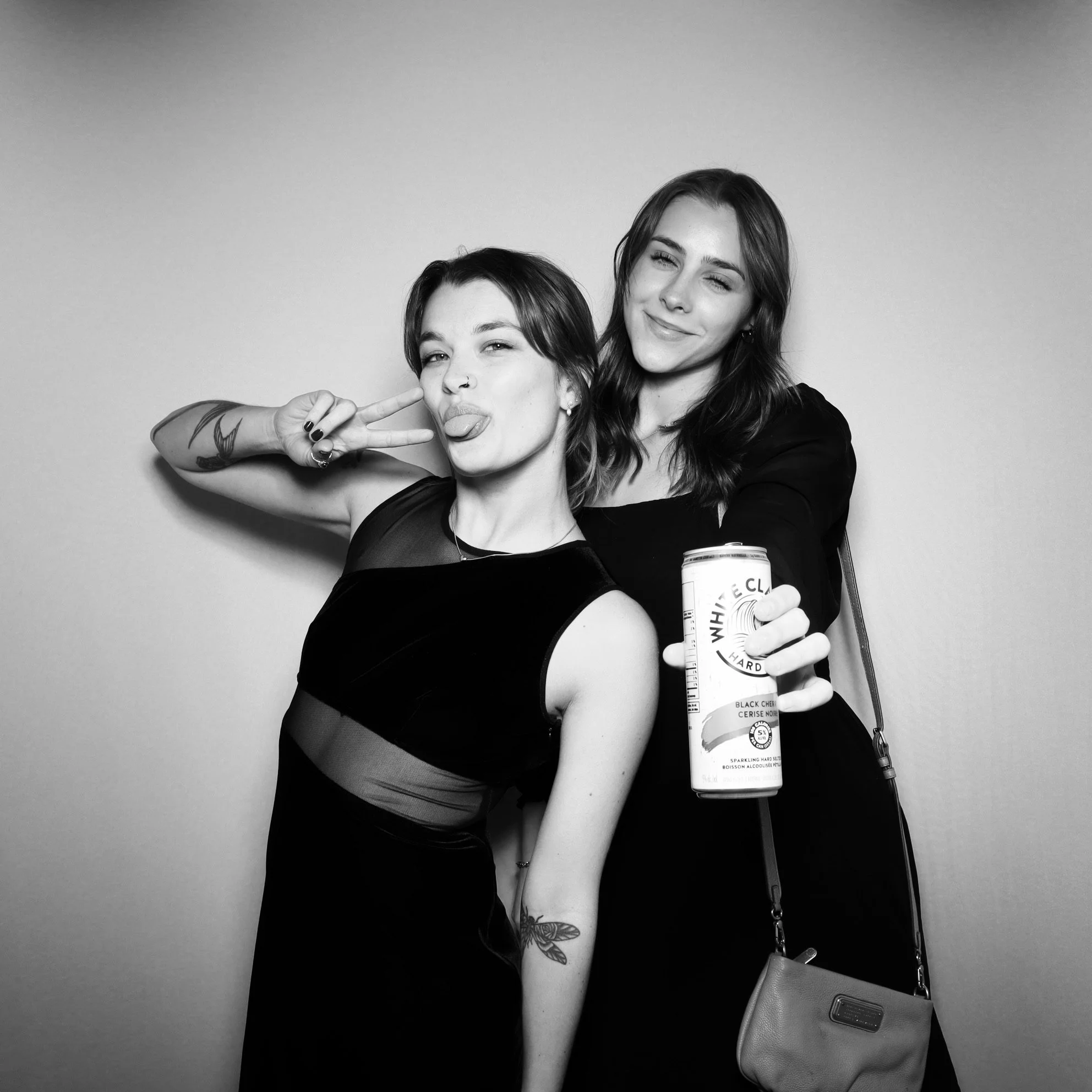 Two young women posing together, one making a peace sign and sticking out her tongue, the other holding a can of White Claw hard seltzer, both smiling and making playful expressions in front of a plain wall, in black and white.